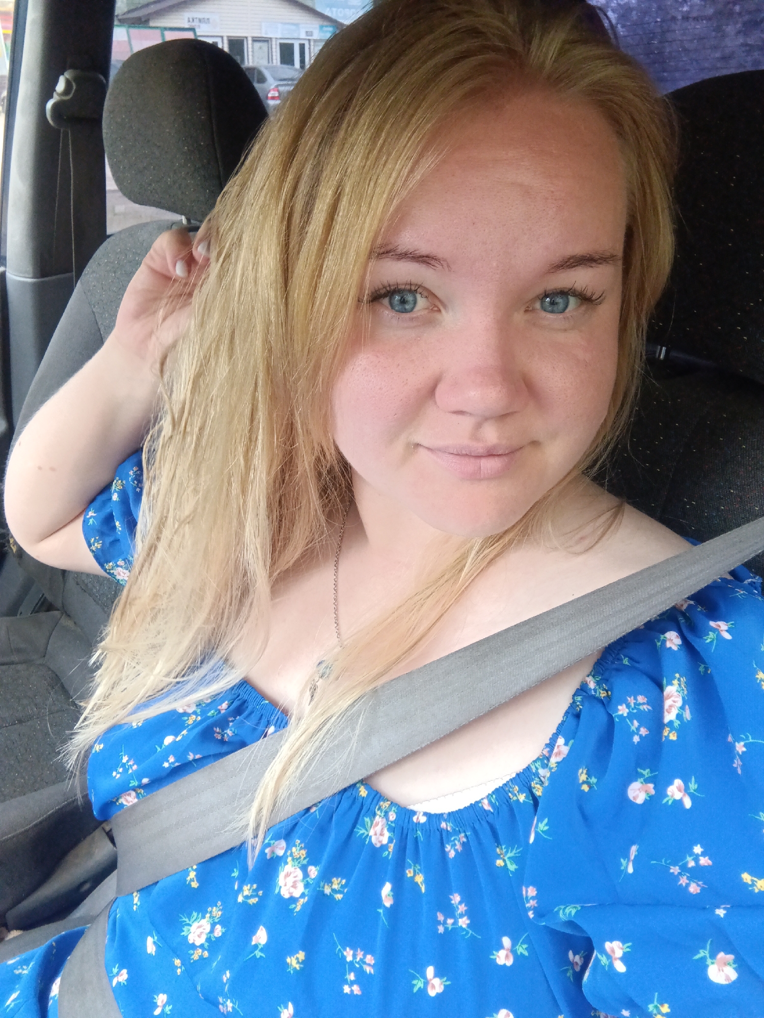 A young Caucasian woman with blonde hair and blue eyes is seated in the passenger seat of a vehicle, wearing a blue floral-print dress and a grey seatbelt. She is looking towards the camera, with her right hand touching her hair. The background shows a blurry view of buildings and cars, suggesting an outdoor setting likely within a city environment. The scene captures a moment of casual travel.