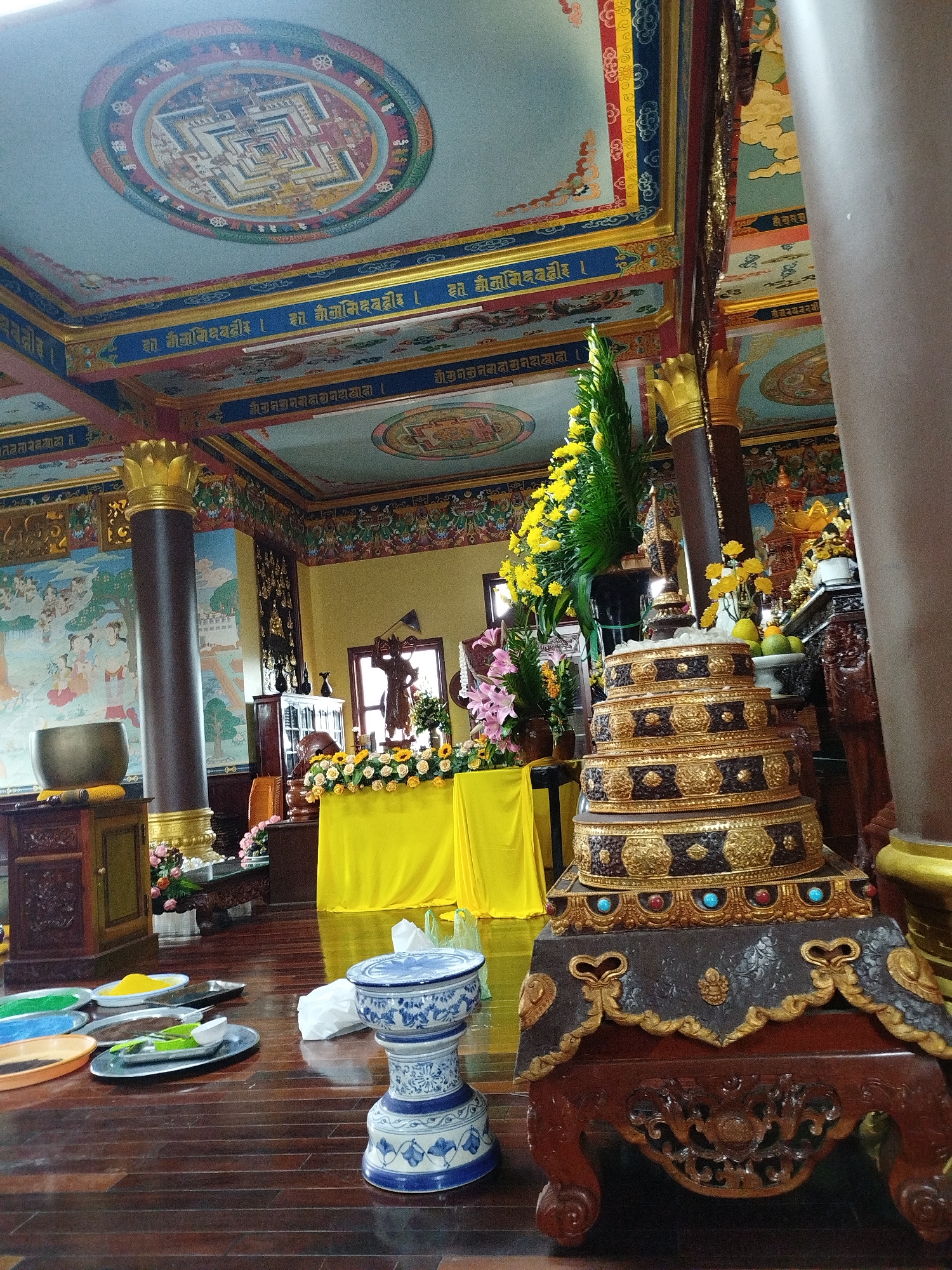 The image depicts the interior of a Buddhist temple, likely in Da Lat, Vietnam, judging by the architectural style and vibrant, colorful decor often found in Southeast Asian temples. The scene is richly adorned, suggesting a place of worship or spiritual significance.
Dominating the foreground and right side of the image is a multi-tiered, ornate golden structure, possibly a stupa or a reliquary, resting on an intricately carved wooden base. The structure is layered with dark brown and gold decorative elements, some inlaid with what appear to be blue and red stones. To the left, a large, dark brown pillar, topped with a gold lotus-like capital, stands prominently. Beside it, a metal bowl, perhaps a singing bowl, rests on a wooden cabinet.
The background reveals a vibrant scene. A yellow altar cloth covers a surface, behind which a statue of a standing Buddha figure is partially visible. Numerous yellow and white flowers are arranged on the altar, with greenery interspersed. Further back, a window lets in some light.
The ceiling is a key feature, painted with elaborate mandalas in circular frames and adorned with decorative borders featuring golden calligraphy and cloud motifs. The walls are also decorated with murals depicting figures and scenes.
On the wooden floor in the lower left, several shallow plates contain colored powders, including yellow, blue, and green, suggesting offerings or ritualistic materials. A blue and white porcelain pedestal sits on the floor near the center-right. The wooden floor itself is polished and reflects some of the light.
The overall impression is one of a well-maintained and richly decorated religious space, filled with symbolic elements and vibrant colors, creating an atmosphere of reverence and spiritual activity.