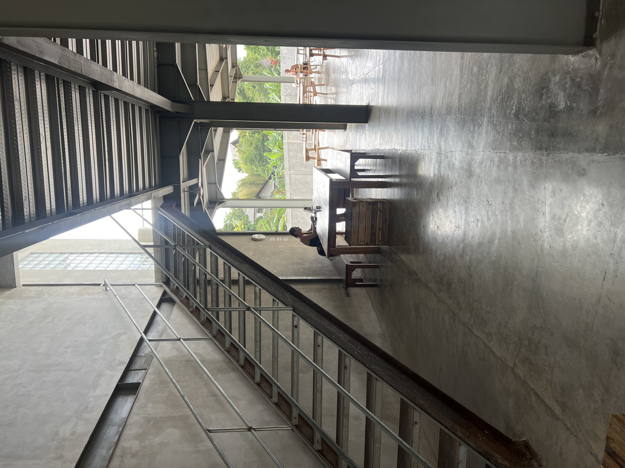 The image depicts an indoor/outdoor cafe space in Mengwi, Indonesia. A metal staircase with wooden steps and metal railings ascends on the left side of the frame. The ground floor area is paved with concrete. In the foreground, a wooden table with a bench is set up, with a person sitting at the table, looking at a laptop. Behind this table, to the right, another wooden table with chairs is visible. Further into the background, more outdoor seating areas with wooden tables and benches are arranged. Lush green trees and vegetation are visible beyond the seating area, suggesting an open-air environment. The architecture appears to be industrial with exposed metal beams and concrete walls. The lighting suggests it is daytime, with natural light filtering in from the open sides and a window composed of glass blocks high up on the left wall.