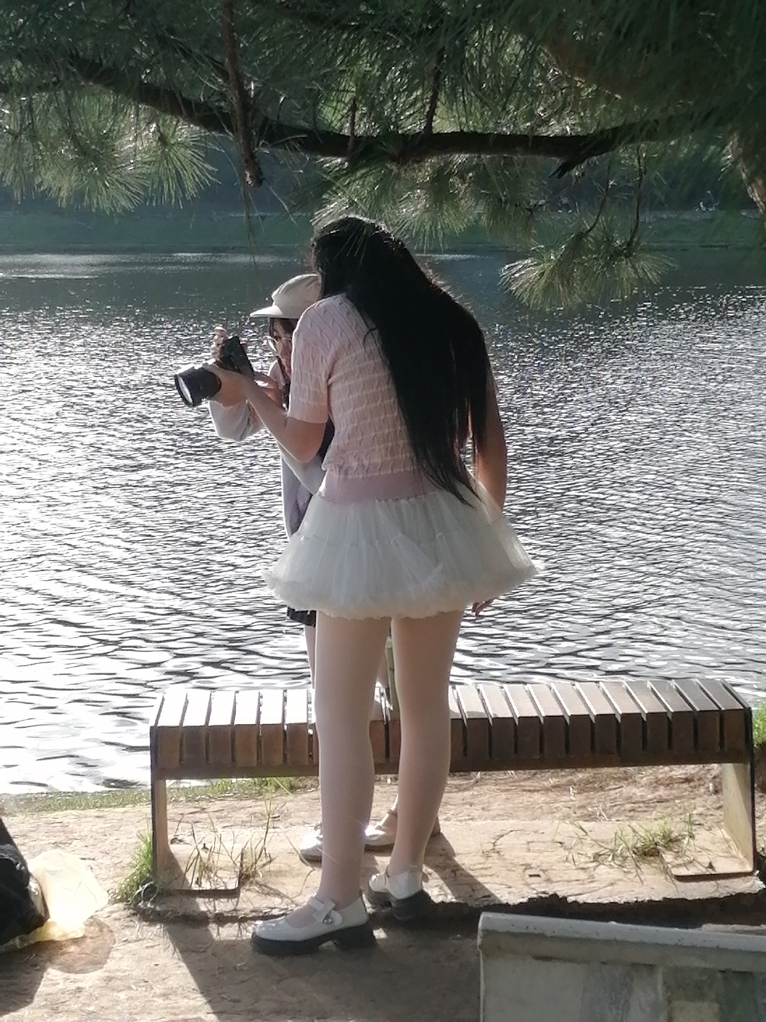 The image depicts two individuals near a body of water, likely a lake, in an outdoor setting. The person in the foreground, viewed from behind, is wearing a pink knitted top with a white ruffled skirt and white Mary Jane style shoes. They have long, dark hair. This person is holding a camera, appearing to take a photograph. Behind them, another person is partially visible, wearing a white cap and glasses, also seemingly engaged with the camera. A wooden bench with a slatted design is situated in the middle ground, and the surface around the individuals appears to be a mix of dirt and grass. The water exhibits ripples and reflections, suggesting it is a calm day. In the background, trees and foliage are visible, with a hint of what could be a distant shoreline. The overall lighting suggests it is daytime, possibly late afternoon, given the shadows.
While the image itself doesn't explicitly state the location, the description of the environment – a lake surrounded by pine trees and a somewhat temperate climate – is consistent with areas around Da Lat, Vietnam. Da Lat is known for its lakes, pine forests, and cooler weather, making it a plausible setting for this scene.