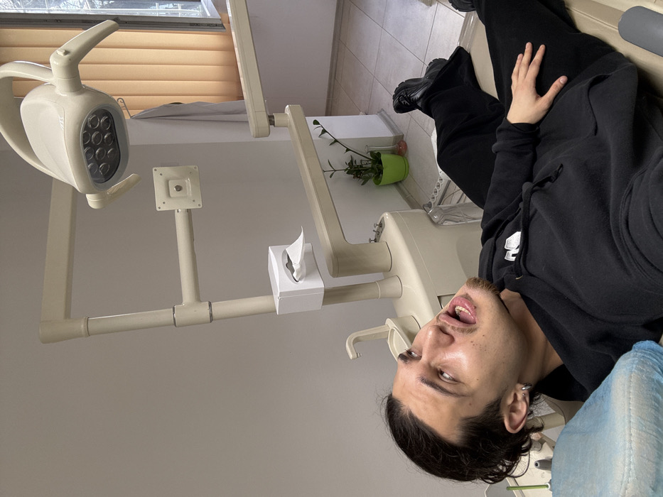 The image depicts the interior of a dental examination room located in Sofia, Bulgaria. A male individual with dark hair, a mustache, and a goatee is positioned on his back in a light-colored dental chair, which has a blue disposable headrest cover. He is wearing a black hooded sweatshirt, black pants, and black shoes. His head is tilted back, eyes are rolled upwards, and his mouth is wide open, exposing his teeth, conveying a state of discomfort or during a medical procedure. His left hand rests on his stomach.
Adjacent to the dental chair is a multi-functional dental unit, also light-colored, featuring a small sink/spittoon, a faucet, and an integrated white tissue dispenser with a single tissue exposed. An overhead, articulated dental lamp with multiple LED lights is positioned above the patient. In the background, a light-colored wall is visible, along with a window fitted with beige vertical blinds. A white coat or smock hangs on a hook near the window. A small green plant in a bright green pot is visible on the floor beneath the window. The floor is covered with light-colored tiles.