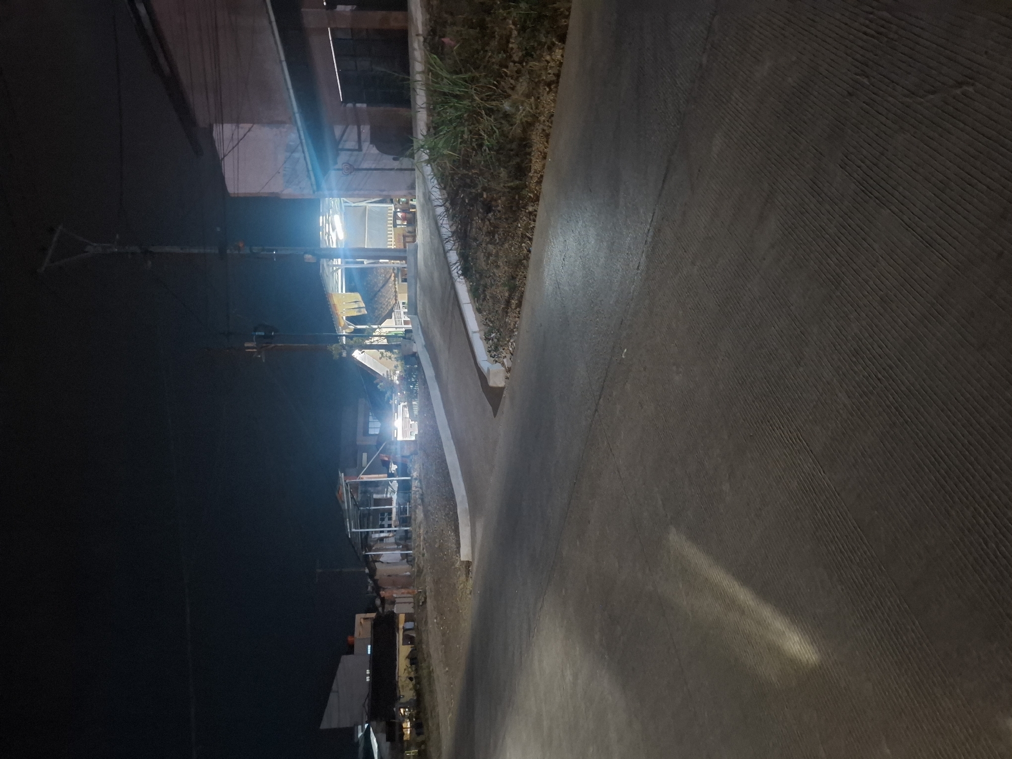 The image depicts a nighttime scene on a paved road in Ciheulang Tonggoh, Indonesia. The road curves from the foreground into the mid-ground, with a section of dirt and dry vegetation on the right. In the background, several buildings with illuminated interiors are visible, suggesting a residential or commercial area. One building has a covered outdoor space with multiple lights. A utility pole with wires stands prominently on the right side of the scene. The sky is dark, indicating it is nighttime. No people are visible in the image.