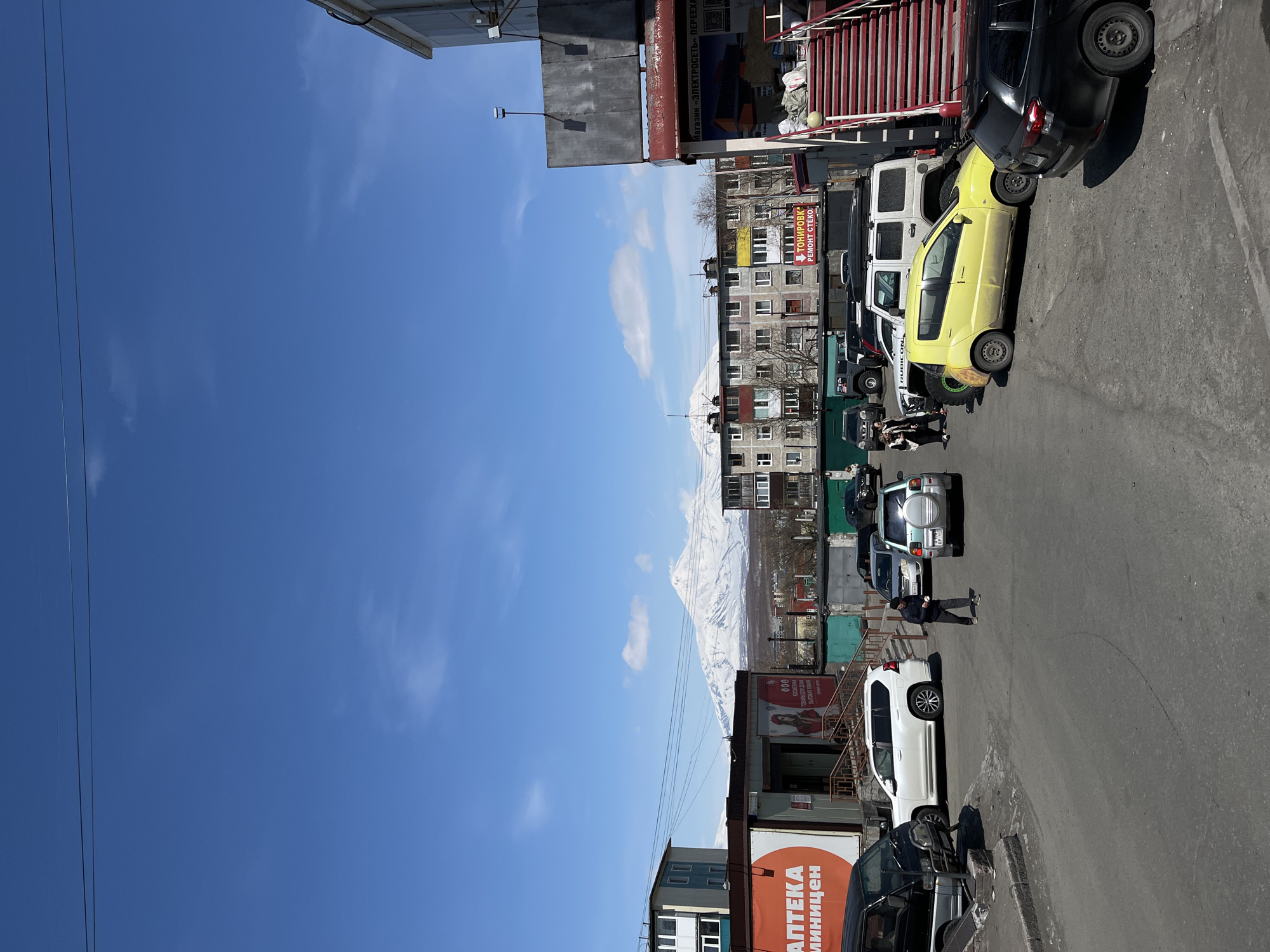 The image depicts a street scene in what appears to be a residential area, with a backdrop of a snow-capped mountain range. The sky is clear blue. Several vehicles are parked along the side of the road, including a yellow car, a white SUV, and other cars of various colors. Buildings line the street, with signs in Russian, including "APTEKA," suggesting a pharmacy, and another sign for "TONIROVKA," likely indicating a car tinting service. A person is seen walking on the street, interacting in the environment. The overall scene conveys a typical day in a Russian town with a striking natural setting.