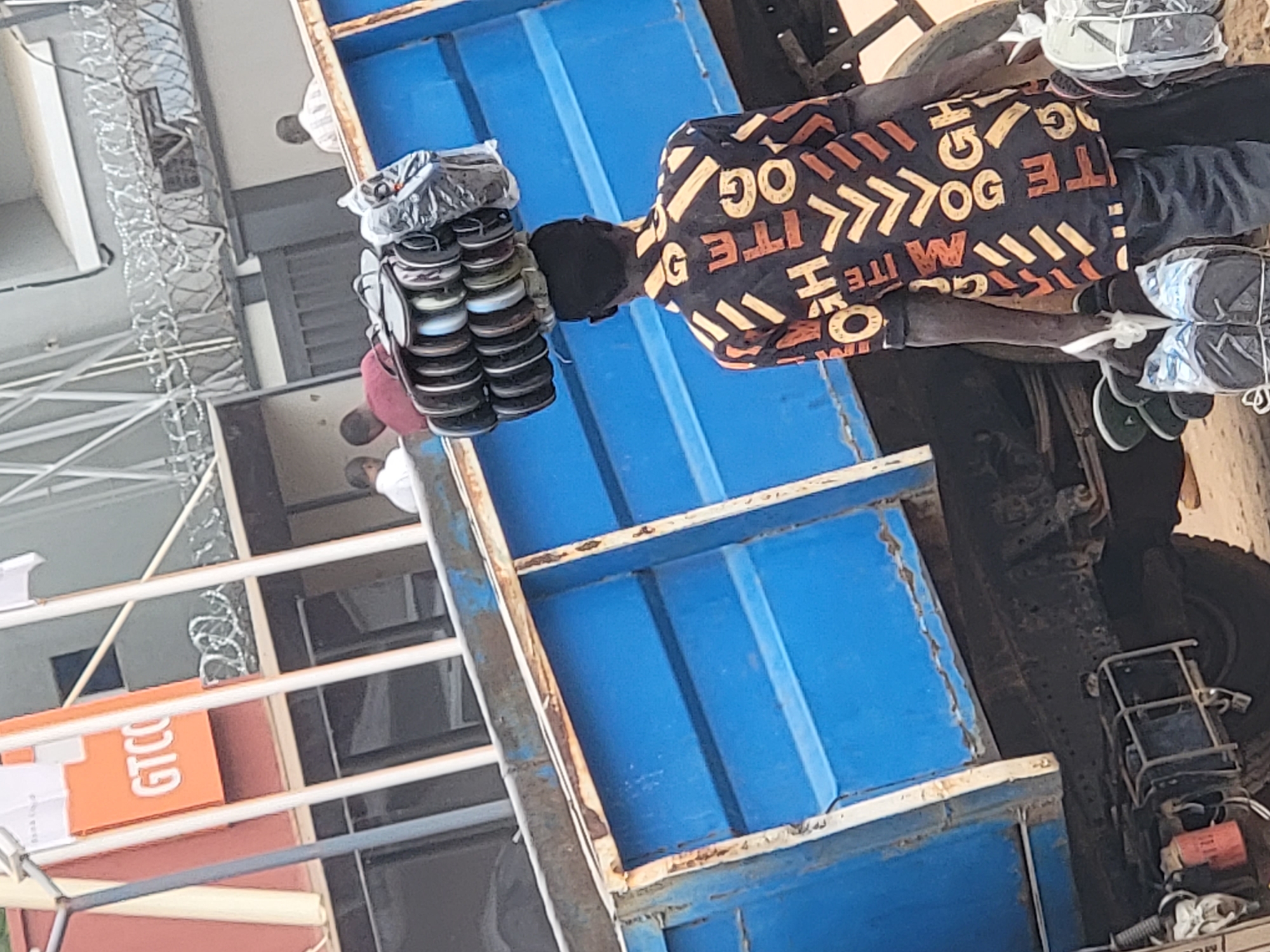 In Maiburiji, Nigeria, a man wearing a black and orange patterned shirt stands next to a blue truck bed holding a stack of flip-flops. Additional stacks of packaged flip-flops are also present. Barbed wire runs above a building with a partially visible "GTCO" sign. Another person stands in the background. The image is angled and appears to be taken from a lower vantage point.