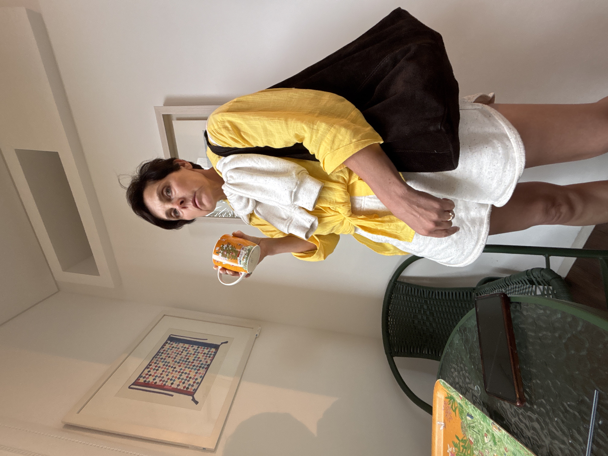 A woman stands in an indoor setting, holding a mug. She is wearing a yellow blouse, white shorts, and a brown tote bag over her left shoulder. A white towel is draped over her shoulders. The room has white walls and a framed picture on the left. A glass-top table with a tray and a dark rectangular object, possibly a phone, sits in the foreground, next to a dark green chair. There is no indication that this scene is located in Seogwipo-si, South Korea.