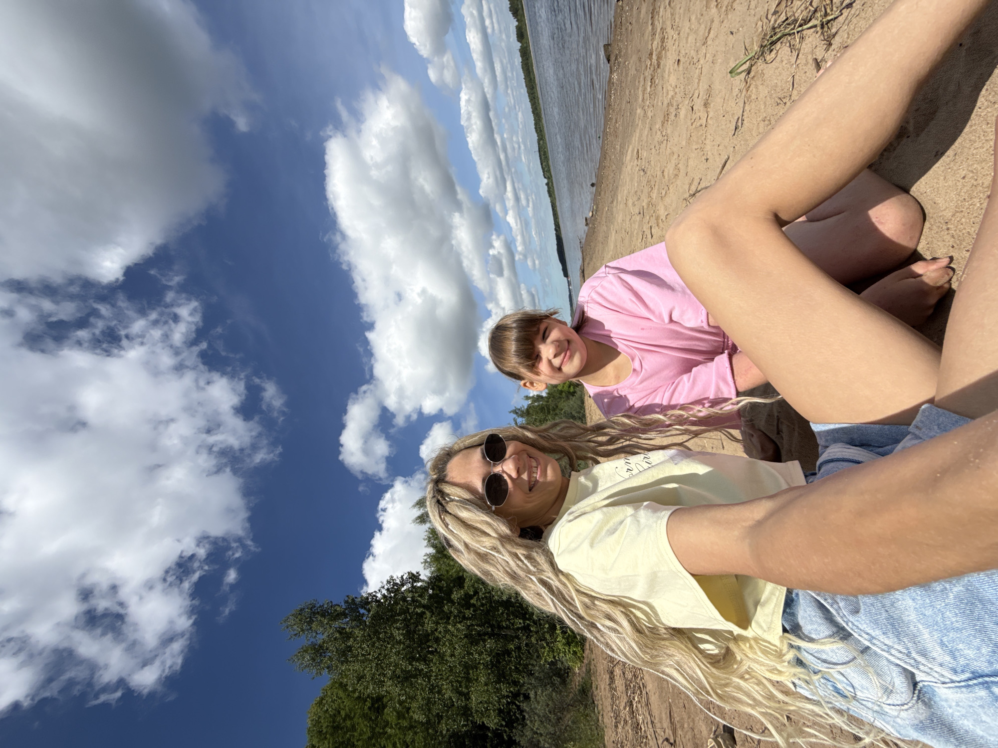 This media file is a selfie taken from a low angle, capturing two individuals on a sandy or dirt bank next to a large body of water, likely a river or lake, under a bright blue sky. The image is rotated 90 degrees clockwise, presenting the sky on the left and the ground on the right.
In the foreground, an adult woman with long, wavy blonde hair wears a light-colored (possibly yellow or cream) t-shirt, denim shorts, and round sunglasses. She is smiling broadly, looking towards the camera. Behind and slightly above her, a young girl with light brown hair and bangs is visible, wearing a pink t-shirt. She also smiles towards the camera. Both subjects appear happy and relaxed.
The setting is outdoors, characterized by a wide expanse of water stretching to a distant bank lined with green trees. The sky is prominent, filled with fluffy white cumulus clouds. The immediate ground where the subjects are seated is dry and sandy. The time of day is clearly daytime, with bright sunlight suggesting good weather. There is faint, illegible cursive text on the woman's light-colored t-shirt. The scene depicts a casual, enjoyable moment by the water, consistent with an outdoor activity in a natural environment like Заволжье, Russia.
