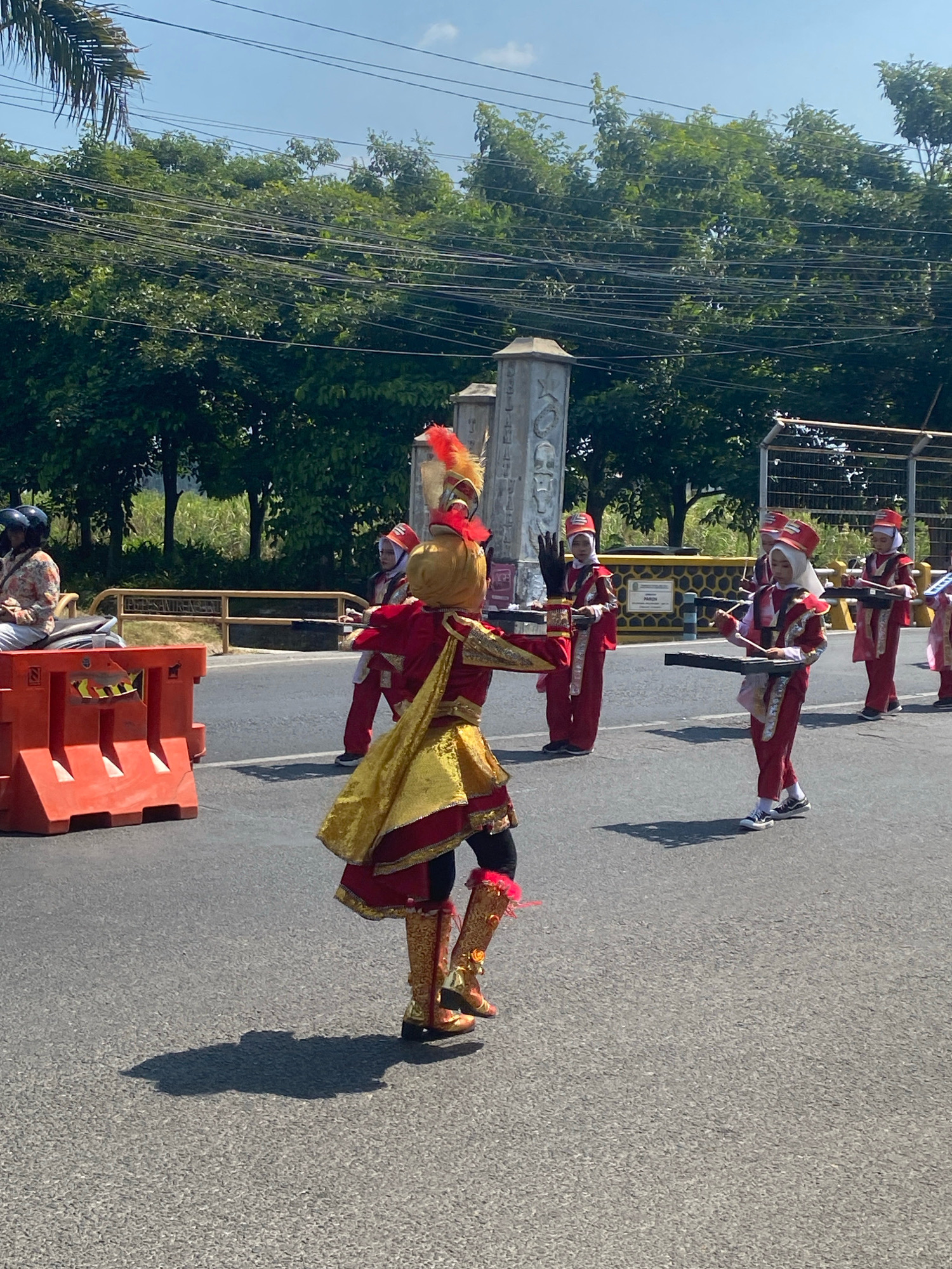 In the city of Ngasem, Indonesia, a marching band procession is underway on a paved road. The participants, dressed in vibrant red and gold uniforms with feathered headdresses, are walking in formation. Many of them are holding black percussion instruments, suggesting they are part of a drum corps or a similar musical ensemble. One individual in the foreground wears an elaborate golden costume with a red and gold feathered headpiece, and appears to be leading or performing a solo. In the background, trees line the roadside, and utility wires are strung overhead. An orange traffic barrier is visible on the left side of the road, and a person on a scooter is partially seen in the distance. The sky is clear and blue, indicating a sunny day.