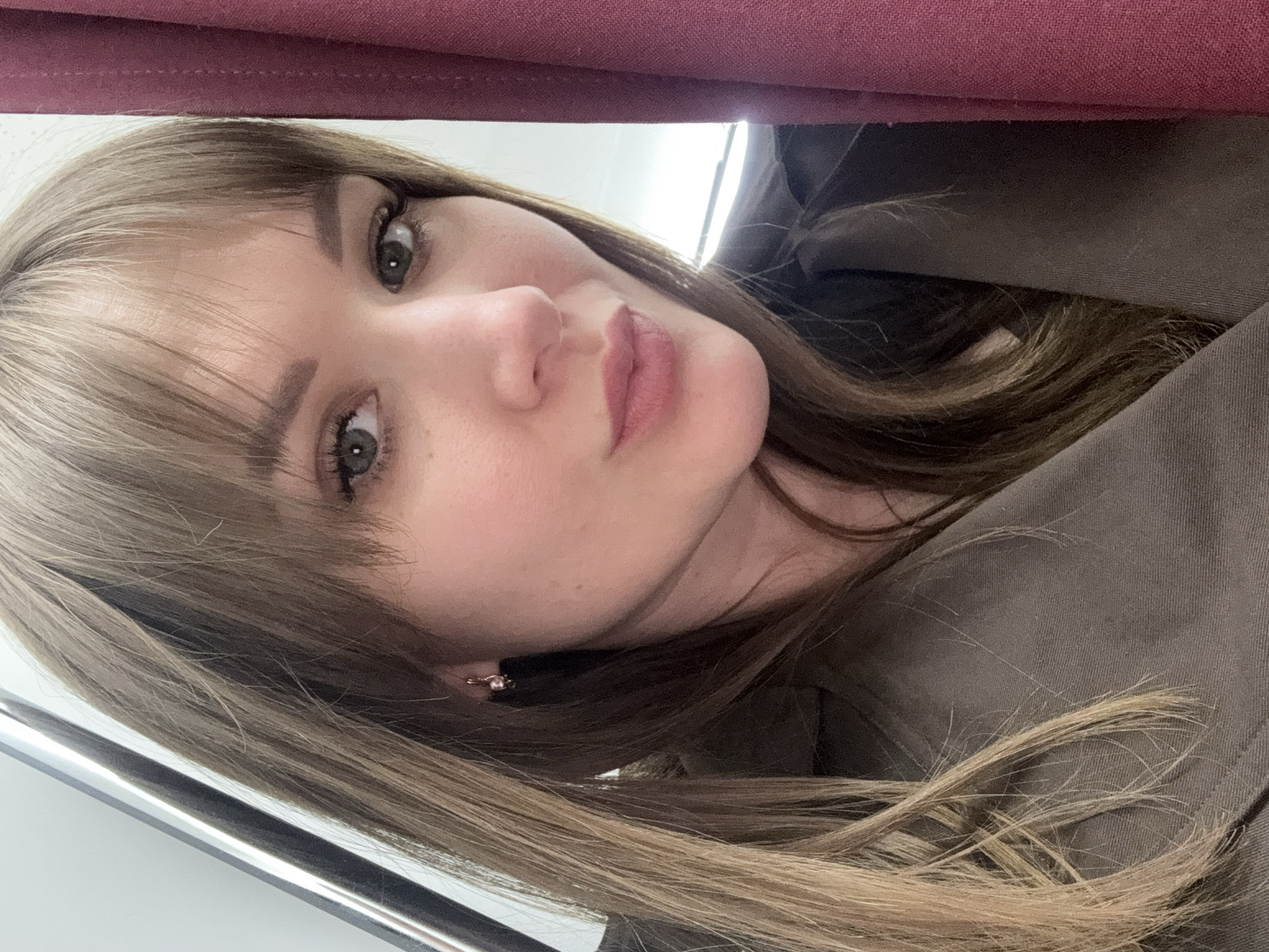 A young woman with light brown hair and bangs is pictured indoors. She is wearing a jacket or coat of a brown-green color. The top of the frame contains a maroon fabric. A thin, silver bar is visible in the bottom left corner. The location is likely Chaikovsky, Russia.