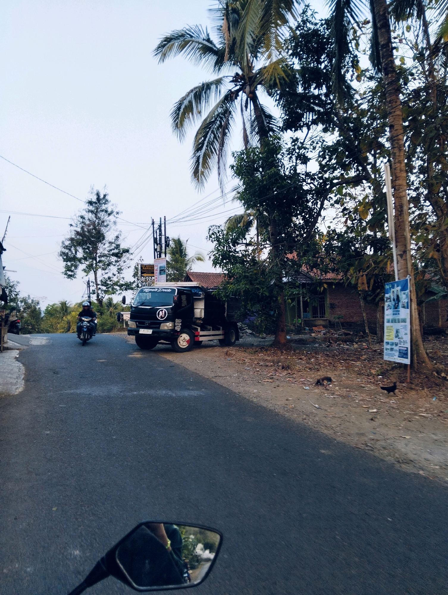 This image captures a street scene in Sidamulih, Indonesia. A black truck with "MAMEN DOCK" written on its side is parked on the right side of the road. A motorcycle ridden by a person wearing a helmet is moving away from the truck on the left side of the road. The road is paved and appears to be in a rural or semi-rural setting with palm trees and other vegetation visible. A brick house with a tiled roof is situated behind the truck. A signpost with posters is also present on the right side of the road. The sky is overcast.