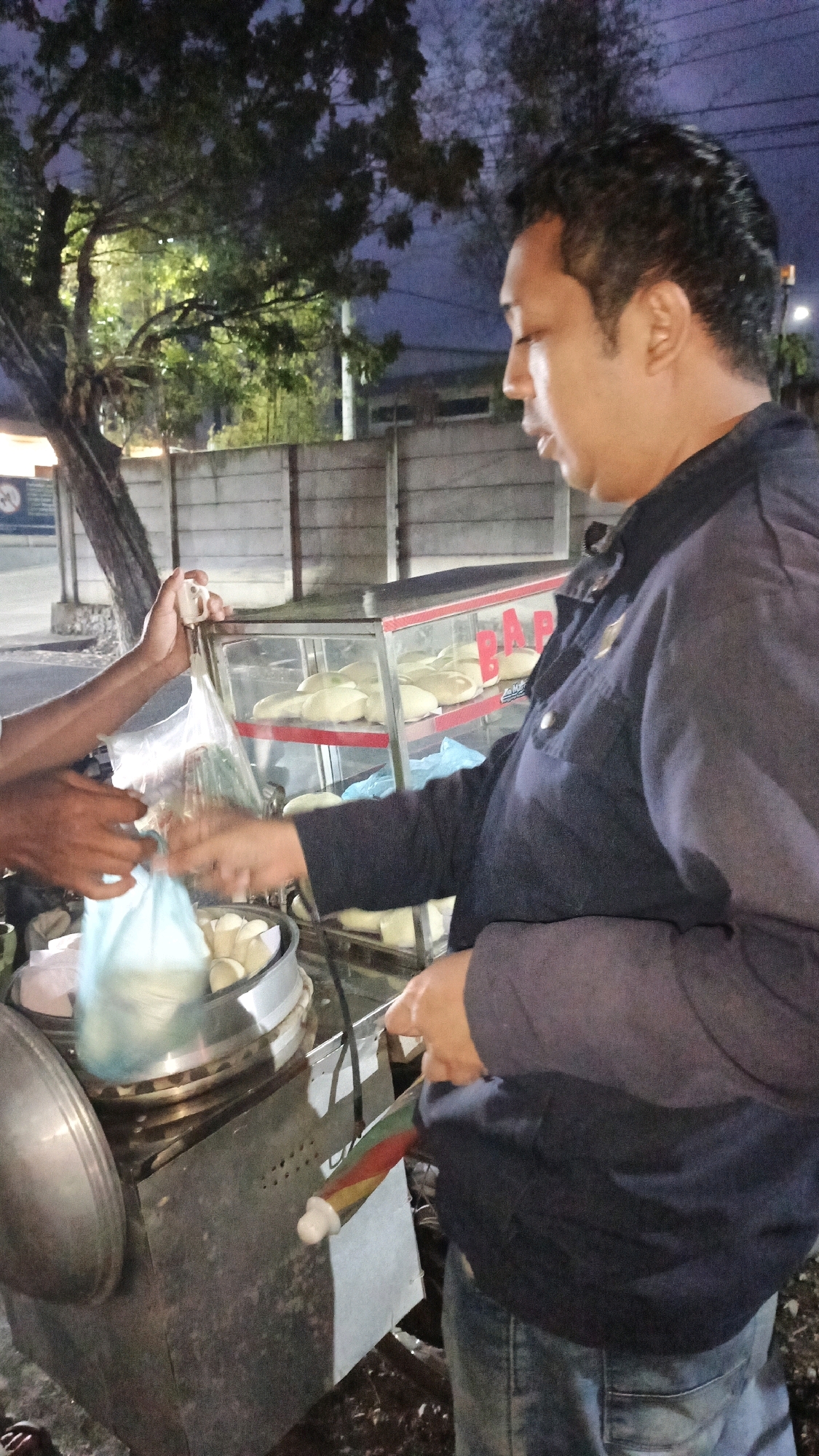 In Sukaresik, Indonesia, at night, a man in a dark jacket and jeans stands by a food cart. The cart has a glass display case filled with white steamed buns. Another person's hands are visible, holding a plastic bag with more buns and placing it into a steamer. The background shows a tree, a concrete wall, and buildings.