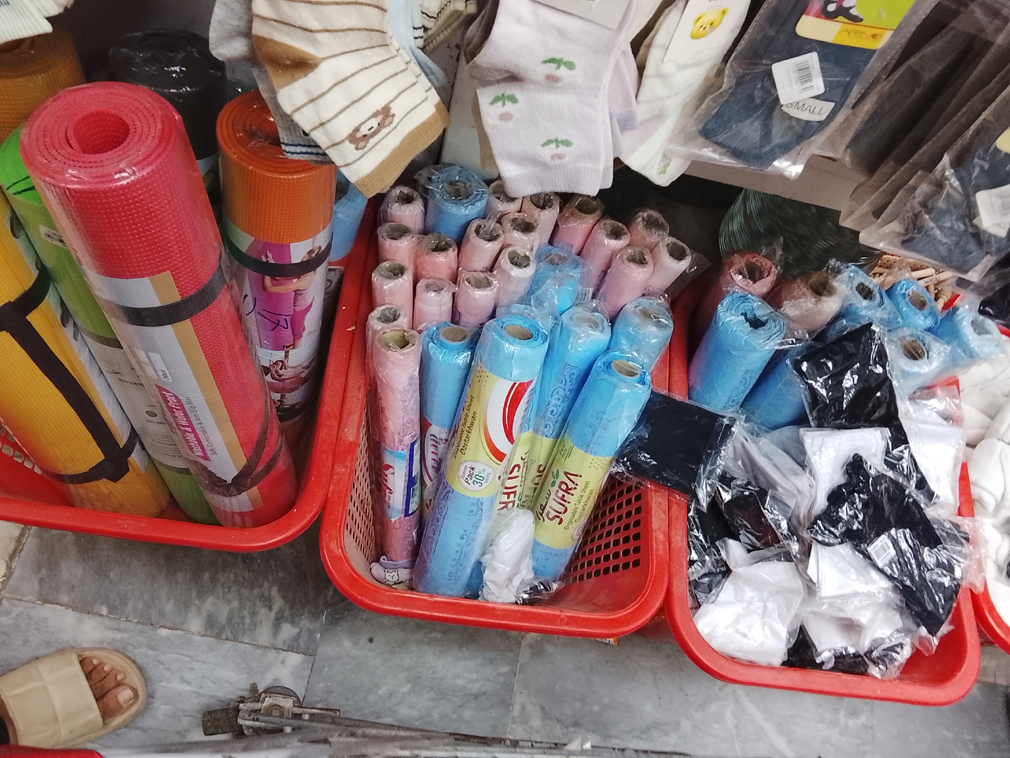 Captured in Gulgasht Colony, Pakistan, the image depicts a vendor's stall or shop corner. Three red plastic baskets form the primary focus. The basket on the left contains rolled-up yoga mats in red, green, and yellow. The central basket holds rolls of plastic tablecloths in shades of pink and blue. One tablecloth brand visible is "SUFRA" Disposable Table Sheet. The right basket contains packages of clothing items, predominantly in black and white. Other items like socks and other clothing articles are visible above the baskets. The floor is grey, and a foot in a sandal is visible at the bottom left. A metal object lays on the floor near the baskets.