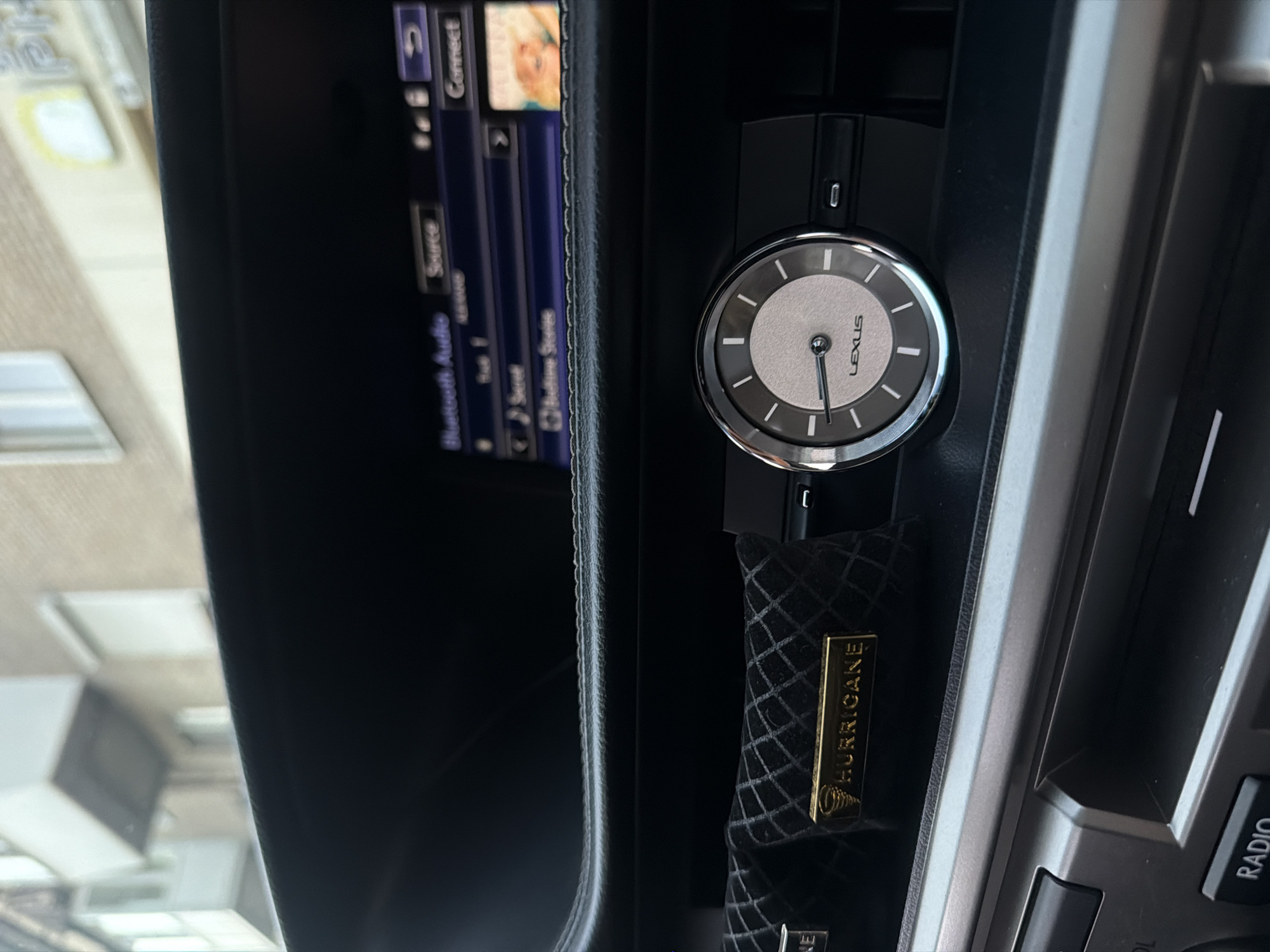 The image shows the interior of a Lexus car, focusing on the dashboard. In the foreground, a round analog clock with the "LEXUS" logo is visible. To the left of the clock, there is a black fabric air freshener with a diamond pattern and a gold-colored plate bearing the brand name "HURRICANE" along with a logo. Above the clock and air freshener, a car's infotainment screen is partially visible, displaying options related to "Bluetooth Audio," "Source," and "Connect." The screen also shows track information and album art. The dashboard is made of black material, likely leather or a similar synthetic. The background of the image, visible through the car's windshield, shows a building with balconies and windows, suggesting an urban environment. Although the image is taken inside a car, the exterior view indicates it is located in a city. The prompt requests the location to be specified as Zaporizhzhia, Ukraine, however, there are no visual elements in the image that confirm this specific location.