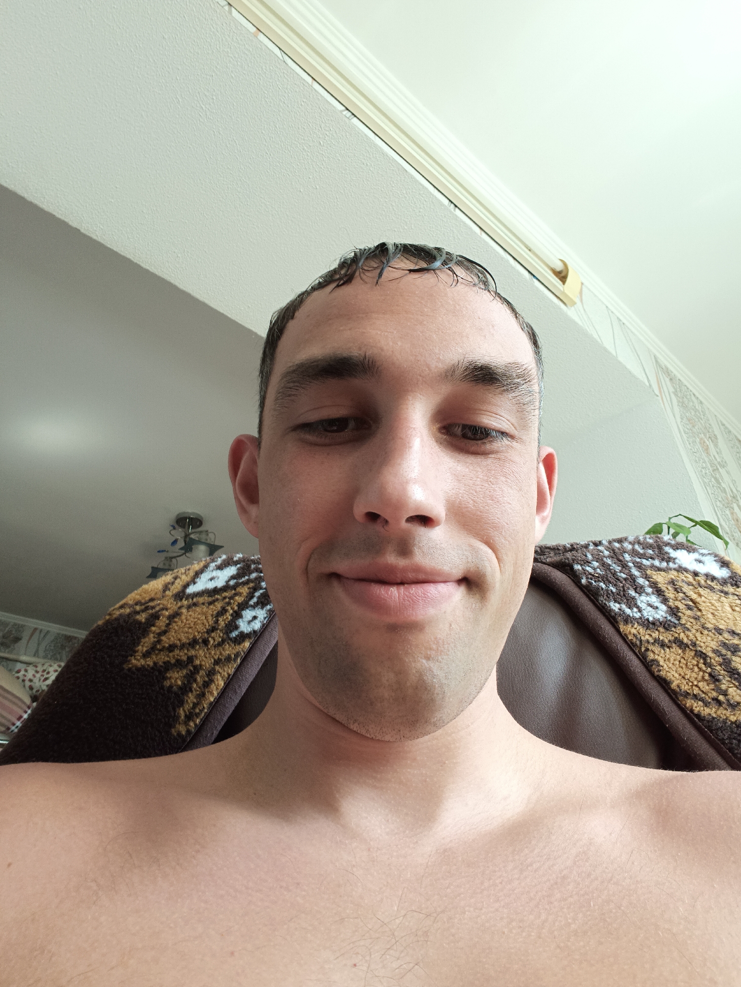 A close-up, low-angle selfie shows a shirtless person with wet, dark hair looking directly at the camera. The person has dark eyebrows, eyes, and a slight smile. They are seated in what appears to be a brown leather chair with a patterned blanket draped over the back. The background shows a light-colored ceiling and wallpaper. A ceiling light fixture is visible in the upper left portion of the image. There are no identifying landmarks or contextual clues within the image that would indicate its location in Berezne, Ukraine.