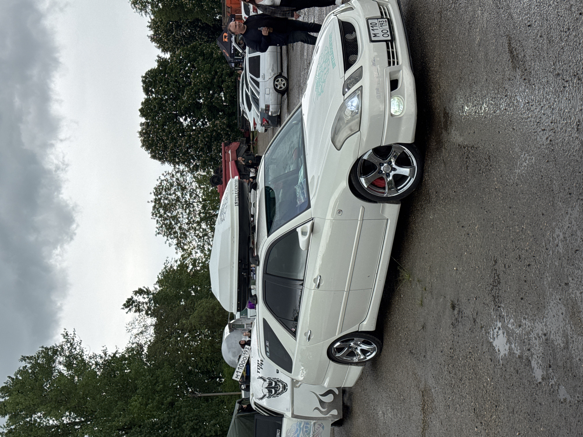A white station wagon with custom wheels and a roof-mounted cargo carrier is parked on a wet asphalt surface. The vehicle features decorative graphics on the rear quarter panel and custom grill work. The license plate reads "M 110 OO 193". Several other vehicles are visible in the background, along with people. The location is Pavlovskaya, Russia, as indicated by the license plate code. Trees are in the background, and the sky is overcast. A man in dark clothing is standing behind the car.