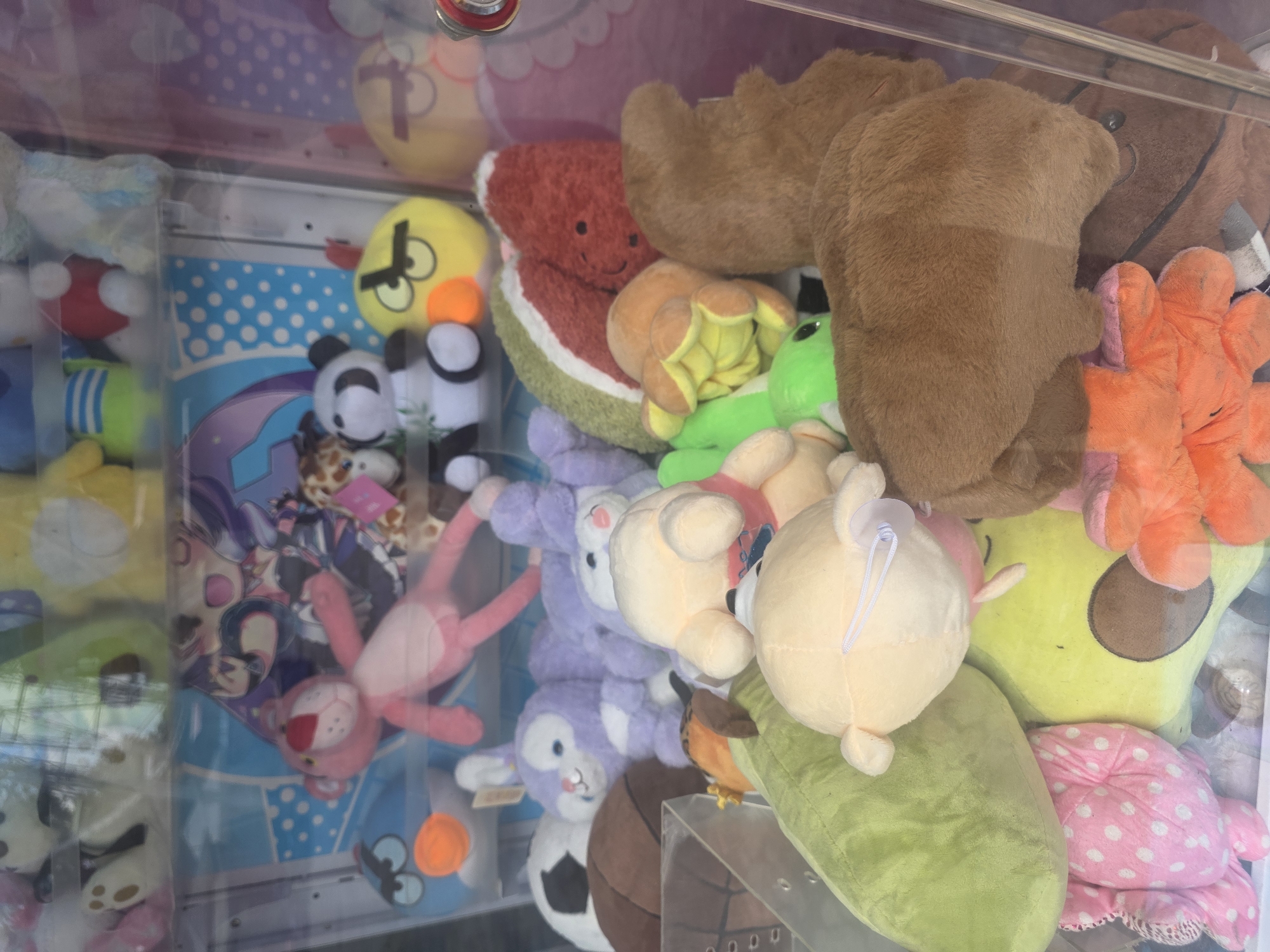 The image displays the interior of a claw machine filled with a variety of stuffed animals. The stuffed animals are of different sizes, colors, and characters, including what appears to be a pink panther, a panda, a yellow bird, and a variety of unidentifiable plush toys. The toys are densely packed within the machine. The claw mechanism of the machine is visible at the top. The background of the machine features a colorful, animated scene with characters and blue and white polka dots. The image is taken from outside the machine, looking in through the glass. There are reflections on the glass, suggesting it is daytime. There are no people visible in the image. There is no information in the image to suggest a location in Bila Tserkva, Ukraine, or any other specific city.