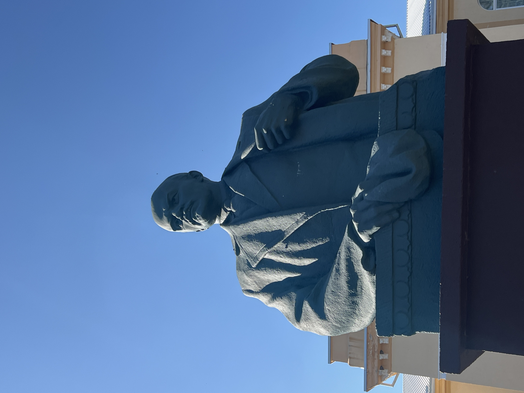 A low-angle medium shot captures a monument of Vladimir Lenin against a clear blue sky. The statue, made of a dark material, depicts Lenin from the chest up, wearing a suit and tie. His right hand is tucked into his suit jacket, while his left arm is bent, with the hand resting on what appears to be a folded garment. The statue stands on a rectangular pedestal adorned with a decorative band. Behind the monument, a light-colored building with white trim and a white roof is visible. The building has architectural details such as decorative moldings and arched windows. The overall impression is one of historical significance, with the statue serving as a focal point in a public space. This monument is located in front of the town council building in Dubăsari, Moldova.