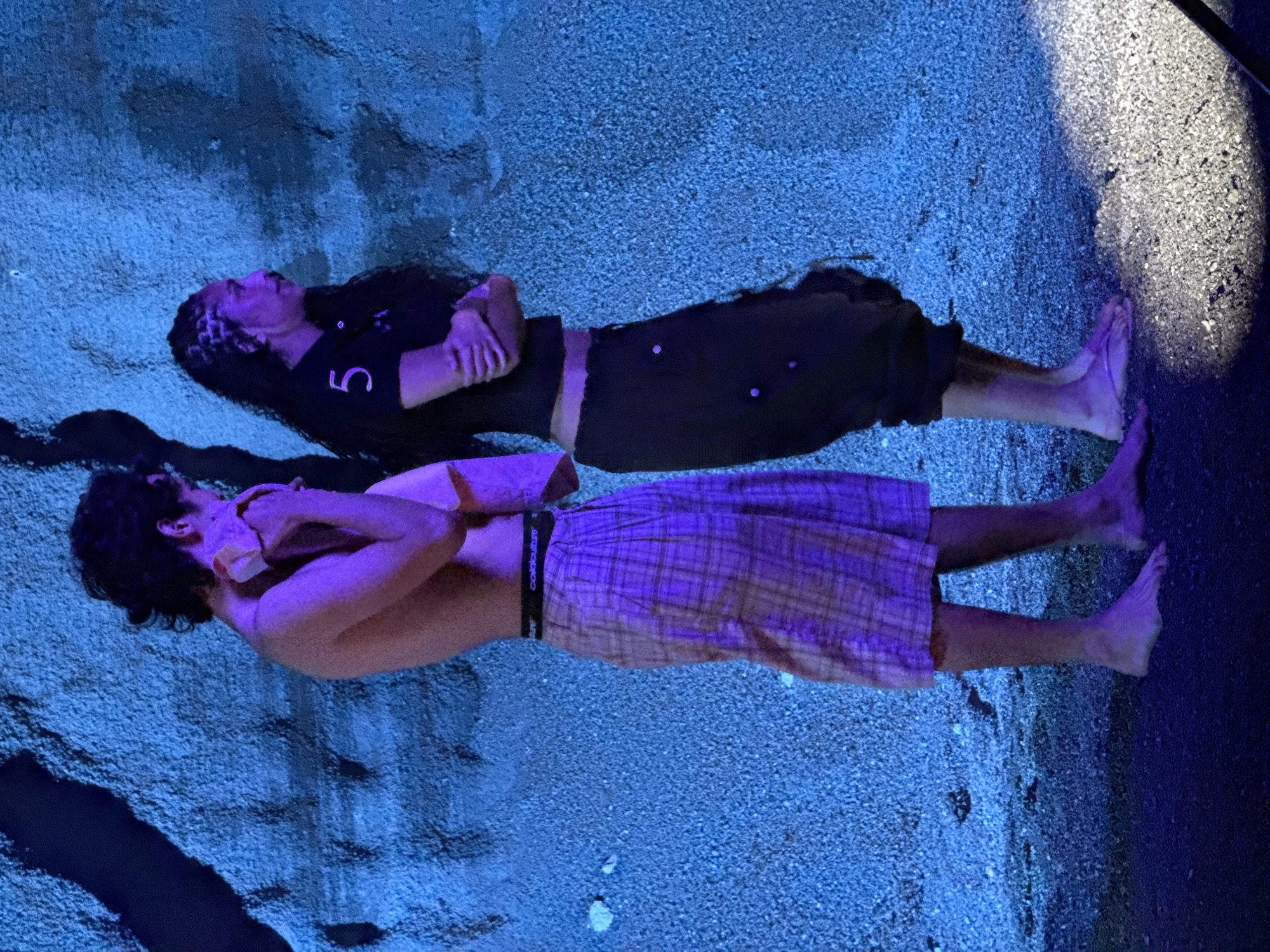 Two people are lying on a rough surface under blue lighting. The person at the top is wearing a black shirt with a white number "5" on it, dark pants, and has long dark hair. The person below them is wearing a plaid skirt, exposing their torso. Both individuals are lying on their backs with their arms crossed over their chests, looking upwards. The surface they are lying on appears to be a rough, textured ground with areas of bright white light and dark shadows. There is nothing that specifically identifies the location as Teguise, Spain.