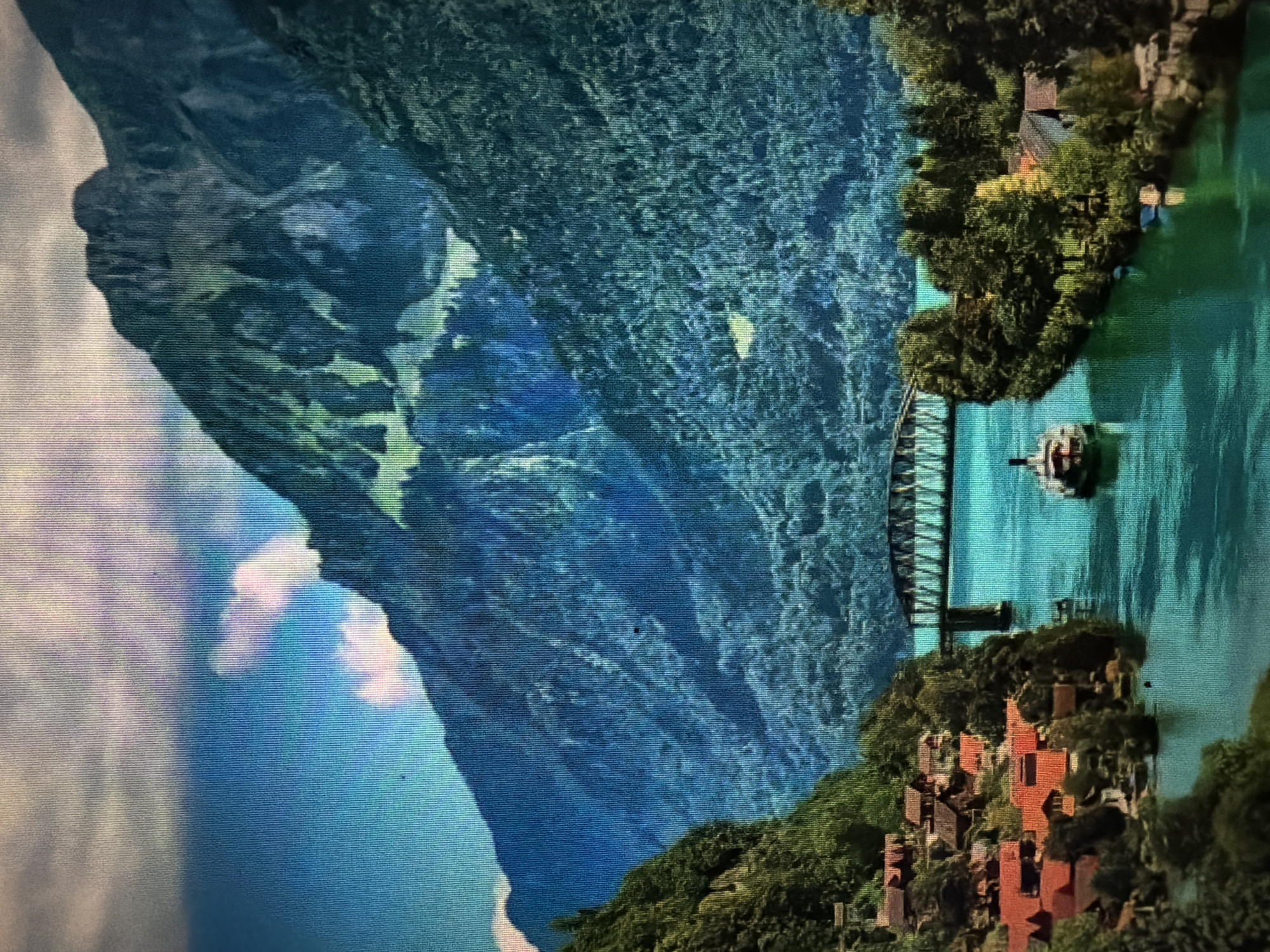 The image depicts a scenic view of a lake or river with steep, forested mountains in the background. A metal truss bridge spans the water, connecting the two sides of the landscape. A small, paddlewheel boat is visible in the water, heading towards the bridge. On the left bank, a cluster of houses with red roofs is nestled into the hillside, surrounded by trees. The water is a vibrant turquoise, and the sky is partly cloudy with patches of blue. There is no discernible indication of the location being the city of Baan Mae Nam, Thailand.