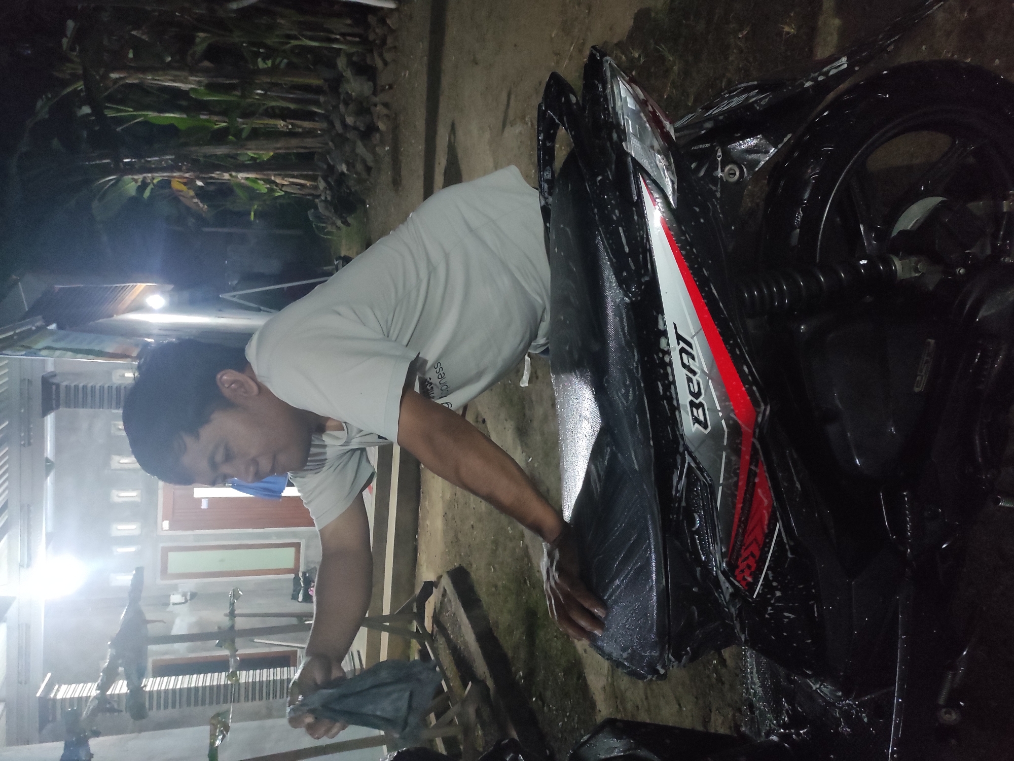 A man is washing a black motorcycle at night in Kemuningsari Lor, Indonesia. The man is wearing a light-colored t-shirt and is leaning over the motorcycle's seat, holding a black cloth in his left hand and placing his right hand on the soapy seat. The motorcycle is partially visible, showing the seat, rear fender, and part of the wheel. The background consists of a building with lights and some vegetation, including banana trees. The ground appears to be dirt or gravel.