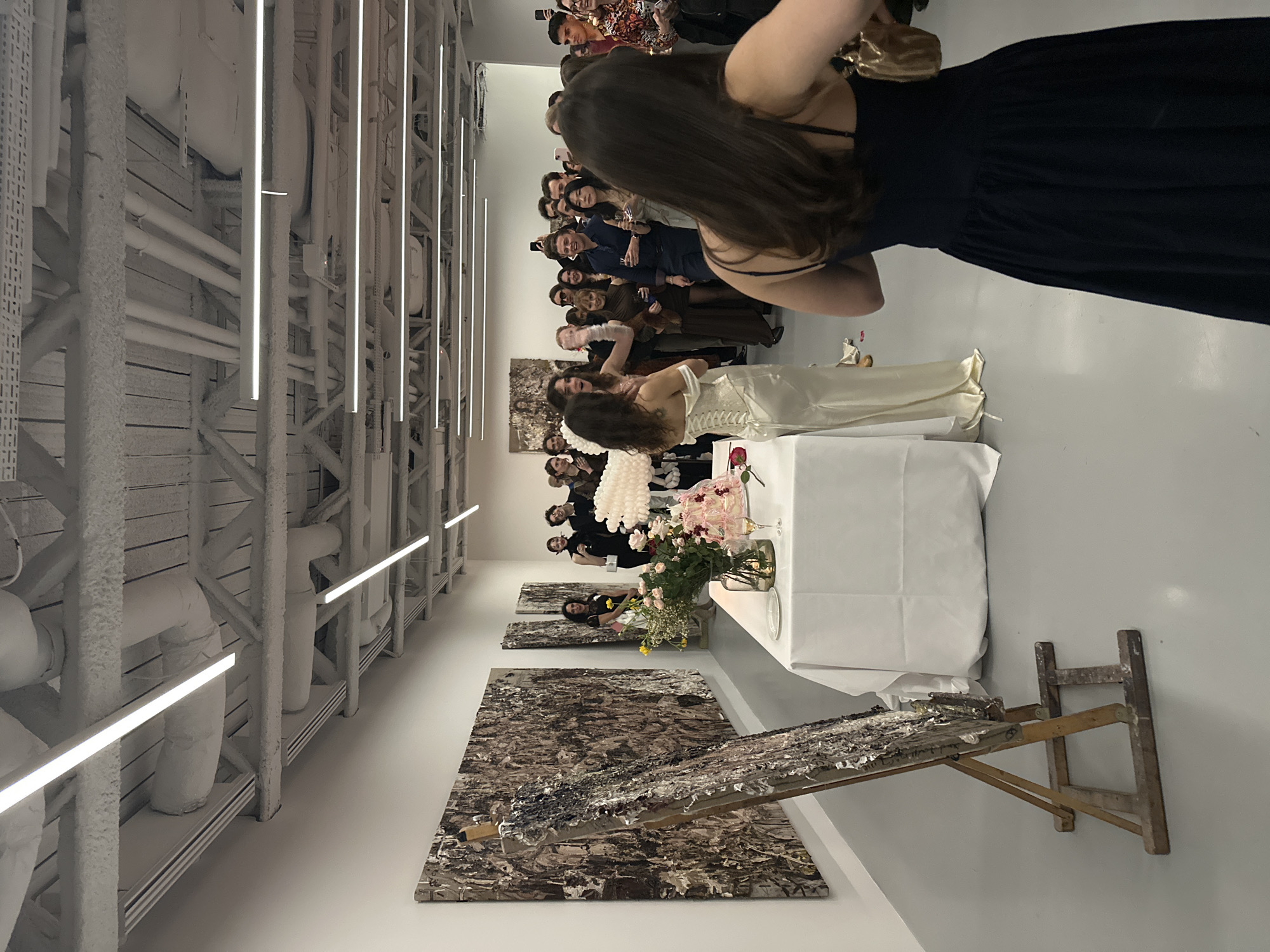 An interior shot depicts an art exhibition or private event within a gallery space located in the City of London, United Kingdom. The room features stark white walls, a light grey floor, and an industrial-style ceiling with exposed white-painted metal trusses, pipes, and ductwork, illuminated by numerous parallel linear LED light fixtures.
In the left foreground, a wooden easel holds a vertically oriented, textured abstract artwork composed of multi-layered, possibly sculptural, elements in grey and black tones. Along the left wall, several large, rectangular abstract artworks with dense, complex textures in earthy brown, grey, and white hues are displayed.
In the mid-ground, a long table draped with a white tablecloth is positioned. It holds a substantial floral arrangement featuring pink and yellow flowers, a smaller vase containing pink roses, a pink-frosted cake or dessert, a champagne flute with a clear liquid, and small white plates.
To the right of the table, a woman in an off-white, corset-style, off-the-shoulder gown stands, facing a gathered crowd. She appears to be interacting with the group, her left arm slightly raised. Partially visible behind her to the left is a large, white, cloud-like sculptural object.
The right side of the image is occupied by a crowd of approximately twenty-five to thirty individuals, both male and female, who are standing and facing the woman in the off-white dress. Many hold clear glasses, consistent with champagne flutes, and some are holding smartphones, suggesting photo or video recording. In the immediate right foreground, the back of a person wearing a dark blue, spaghetti-strap dress and holding a small golden handbag is visible, facing towards the crowd.