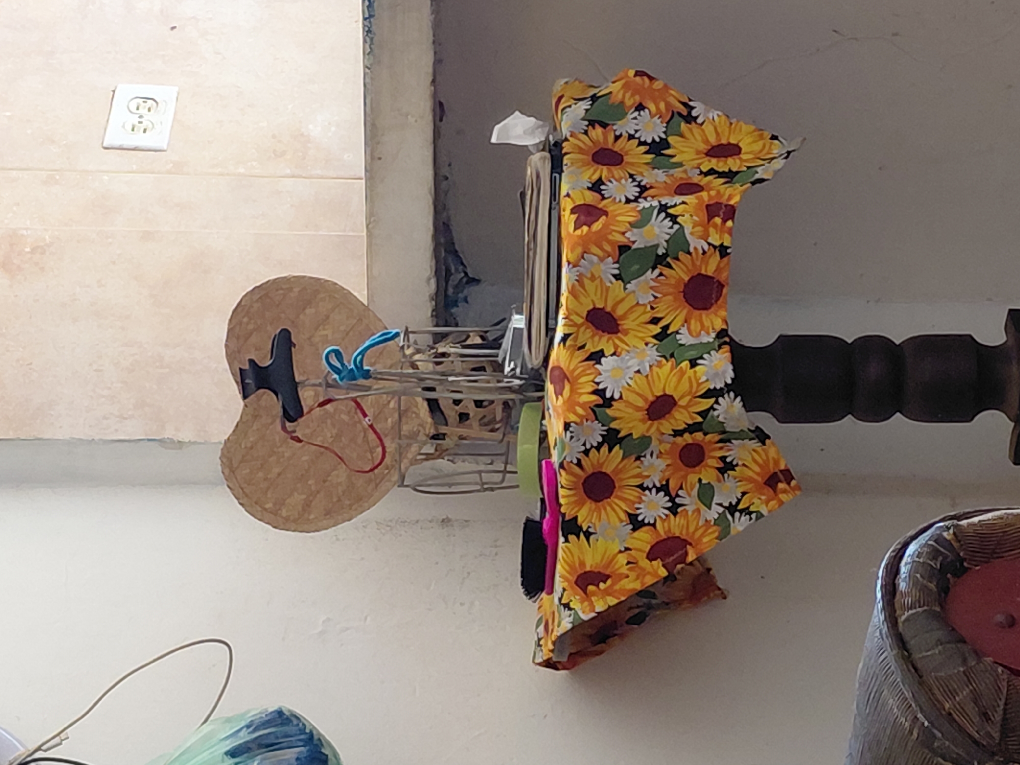 The image shows a small, round table covered with a tablecloth featuring a sunflower pattern. On the table, there are two hairbrushes, one pink and one black, as well as a stack of books or binders and a white tissue. Behind the table, a woven hat with a red cord is displayed, supported by a wire rack. The table itself is dark wood with a carved pedestal base. In the background, a light-colored tiled wall has a white electrical outlet with two sockets. To the left, a plastic bag, possibly containing items, hangs from a wire. The scene appears to be an indoor domestic setting, likely a home or a small shop, possibly in Santa Ana, Venezuela, judging by the style of the electrical outlet and the overall ambiance.