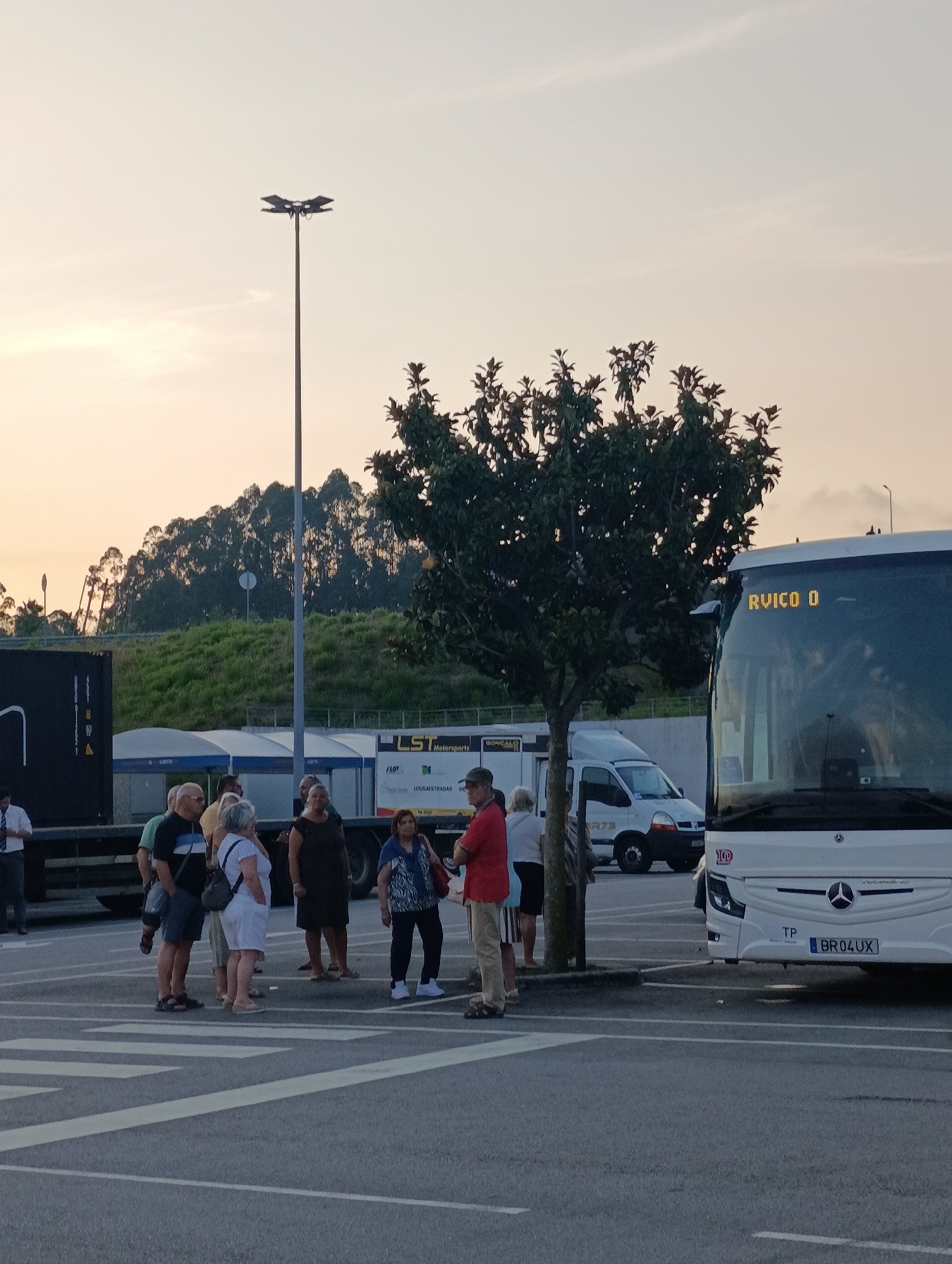 The image depicts a group of people gathered in a parking lot in Santa Maria da Feira, Portugal, near the Europarque. The sky is overcast with a warm, orange hue, suggesting either sunrise or sunset. A large white bus with "RUICO O" displayed on its front is parked to the right of the frame. The bus has a Mercedes-Benz logo and a license plate reading "BR04 UX". To the left, a black shipping container is visible, attached to a truck.
A diverse group of individuals, appearing to be tourists or travelers, are standing around, some engaged in conversation. One woman in a white dress is facing away from the camera, holding a white bag. Another woman in a dark dress is standing beside her. A man in a red shirt and tan pants is also present. A tree with green foliage stands between the group and the bus. In the background, a grassy hillside covered with trees rises, and a tall light pole is visible. Further back, a white van and other structures, possibly part of an event venue or service area, can be seen. The ground is asphalt, marked with pedestrian crossing lines.