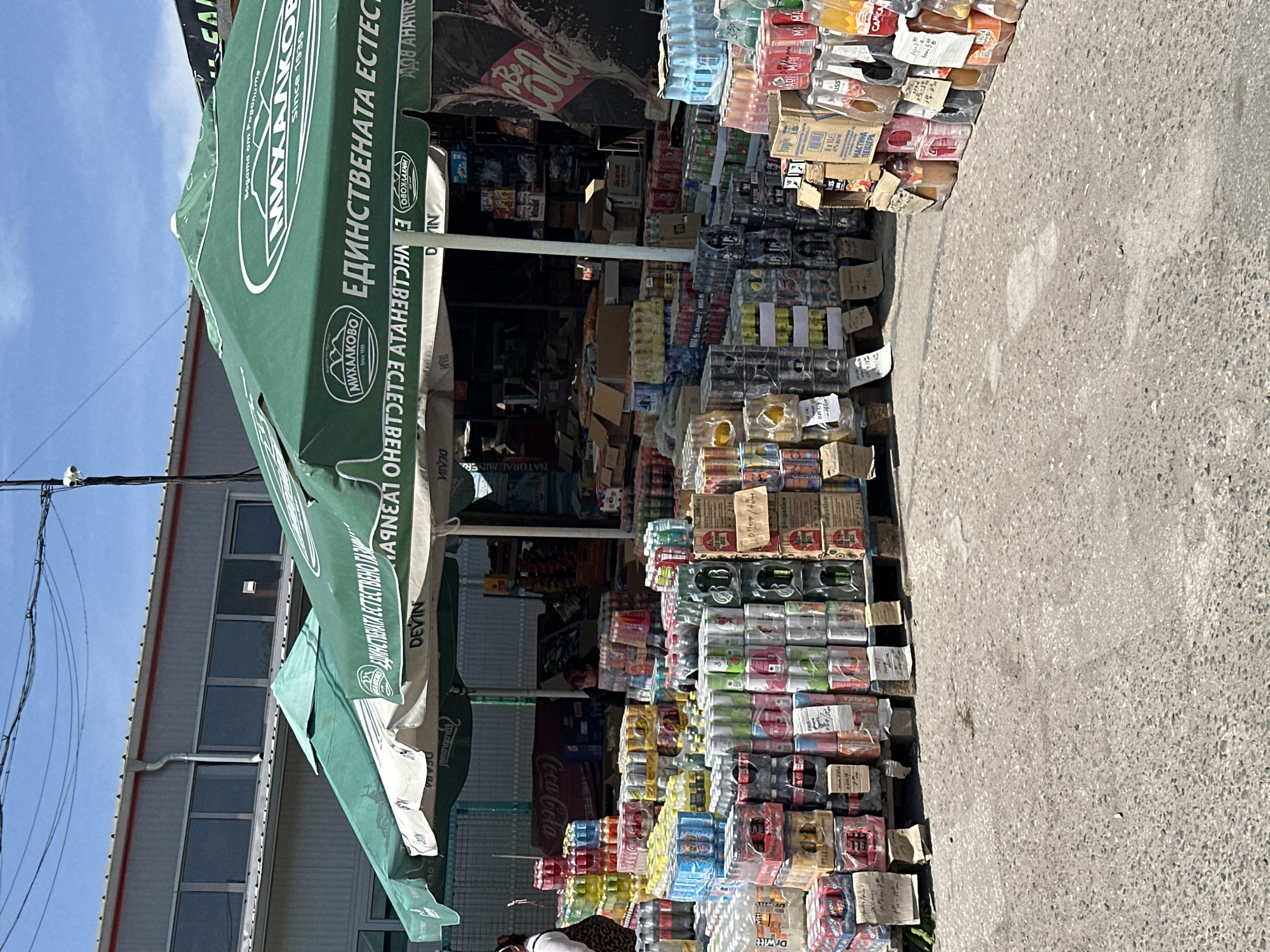 This media file captures an outdoor beverage stall or shop, likely operating roadside in Plovdiv, Bulgaria, given the provided location context and visible Cyrillic text. The scene is bright, indicating daytime under a clear, sunny sky.
The primary subject is a large, informal display of various bottled and canned beverages, stacked high on pallets and cardboard boxes along the edge of an asphalt surface. Products include bottled water, soft drinks, juices, and possibly alcoholic beverages, many with handwritten price tags or labels. Green market umbrellas, one partially collapsed, provide shade over the stall, bearing brand names and slogans. One prominent umbrella features the brand "МИХАЛКОВО" (Mihalkovo) with the text "ЕДИНСТВЕНАТА ЕСТЕСТВЕНО ГАЗЛИКИ" (The only naturally sparkling/gaseous) and "Since 1939", along with "Bogama om Pogonume" (Blessing from Rhodopes).
Other visible brand names and texts include "DEVIN," "Dr Witt," "Coca-Cola" (on a large banner inside the stall), "NATURAL MINERA," "МАСТИКА ПЕЩЕРА" (Mastika Peshchera), "AHAYNE," "uss," "CAMICA," and "BELL CLASSIC BL." The interior of the shop, visible behind the stacks of drinks, appears dim and filled with more products.
A person is partially visible in the lower left corner of the image (when upright), possibly a customer or vendor. Power lines crisscross against the blue sky above a building with dark windows in the background. The overall impression is a bustling, practical outdoor retail setup for drinks.