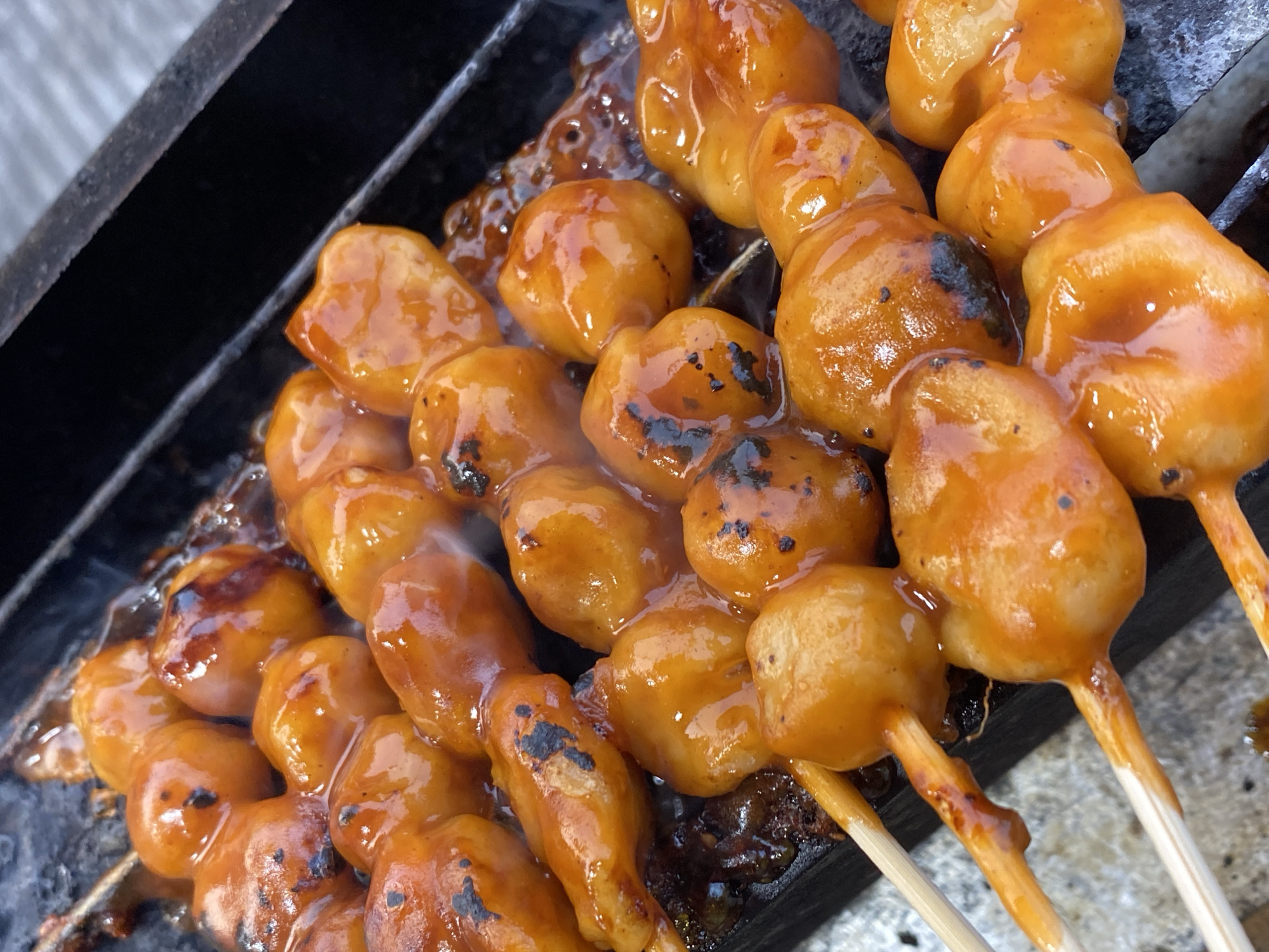 In Kasihan, Indonesia, this close-up image captures several skewers of grilled meatballs or dumplings coated in a glistening, reddish-orange sauce. The food is arranged on a dark, metal grill grate, with some areas showing char marks from the grilling process. Wisps of steam rise from the hot food, suggesting it is freshly prepared. The meatballs appear rounded and slightly irregular in shape, densely packed on thin wooden skewers. The sauce is thick and appears to have a slightly sticky texture, coating each piece and pooling slightly on the grill. The background is out of focus, hinting at an outdoor setting, possibly a street food stall.