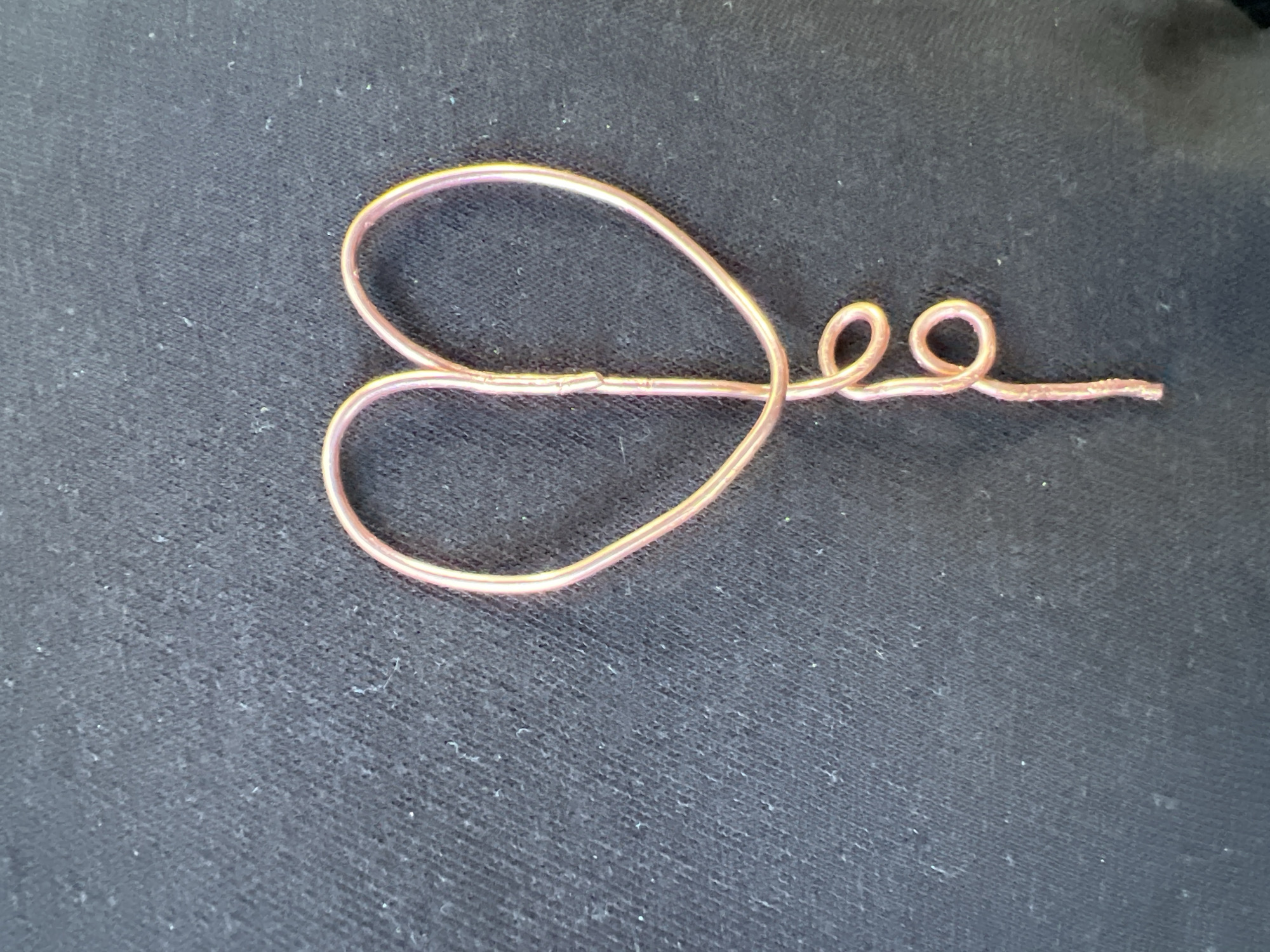 A close-up, top-down shot features a piece of copper-colored wire meticulously bent into an artistic shape, resting on a dark, textured fabric surface. The wire, which appears to have a clear or translucent insulation, reflects light with a soft, coppery-pink sheen. On the left side, it forms a stylized, somewhat asymmetrical heart-like loop. From the point where the heart's two sides meet and cross, the wire extends to the right, creating two distinct, tight spirals before concluding in a straight, unbent segment. The background is a dark, possibly black or very dark gray, fabric with a visible woven texture and scattered specks of lint or small fibers. No people, activities, or text are visible in the image. The lighting is even, suggesting an indoor setting.