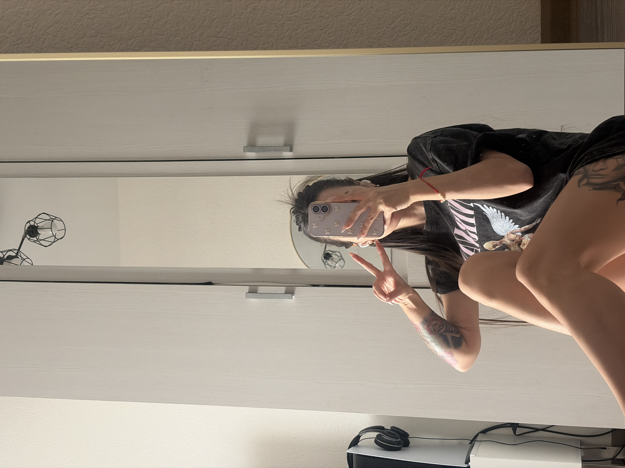A selfie is reflected in a mirror or a mirrored wardrobe door. The person is wearing a black t-shirt and shorts and holding a cell phone with a light purple case. They are making a peace sign with their left hand. The room is mostly white with a wooden border at the top of the image. Beneath the wardrobe, there is a lamp, a PlayStation 5, and headphones. The location is not discernable from this image.