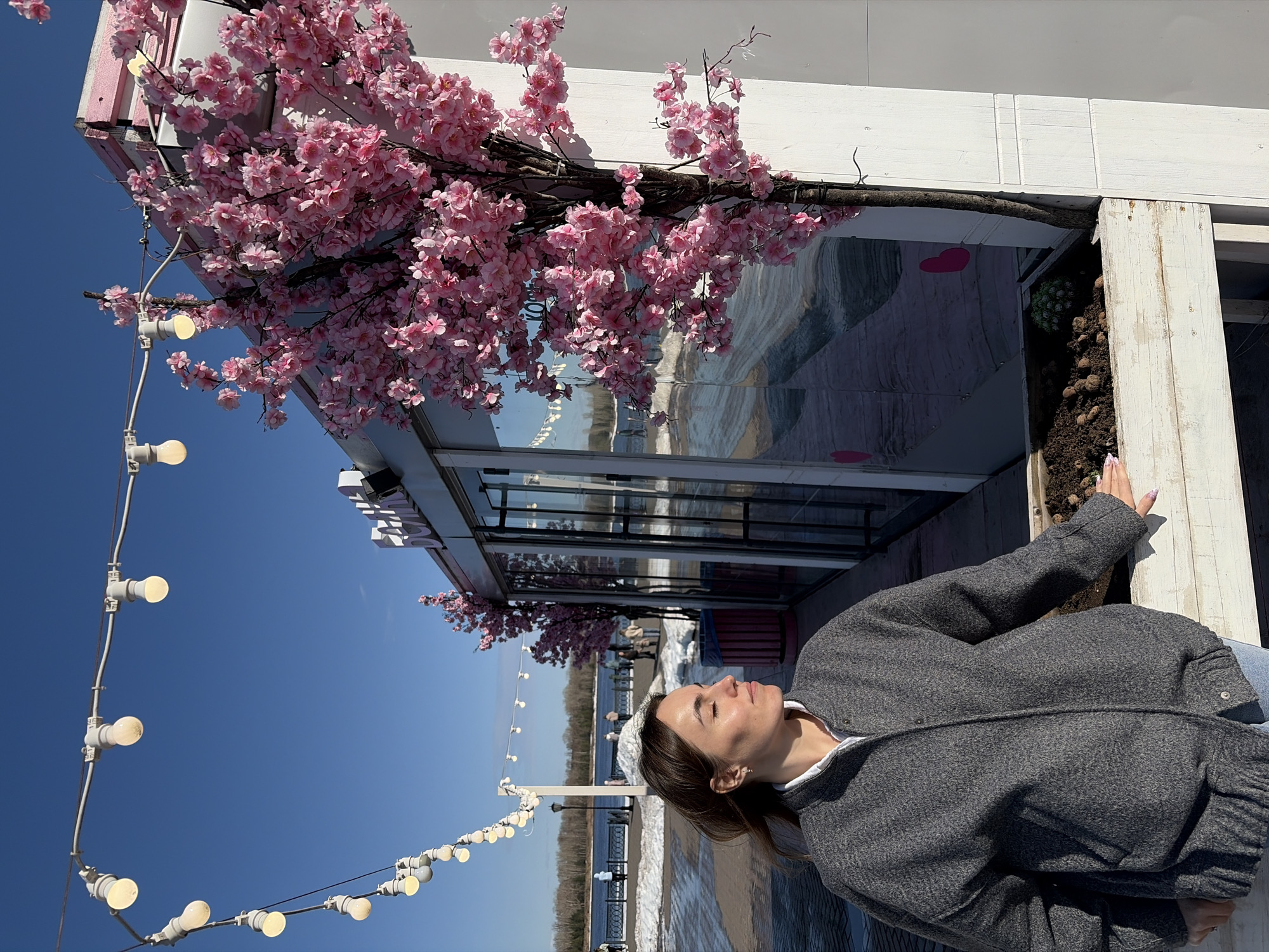 A medium-shot, candid image shows a woman positioned outdoors, looking upward with her eyes closed. She wears a gray jacket over a white collared shirt. A strand of incandescent light bulbs is visible above and to the left. Pink cherry blossoms are situated near the upper left corner, connected to what seems to be a building, as there are windows visible. The location, as specified, is Sarapul, Russia. In the background, a river or body of water is apparent, with trees on the far bank. The sky is clear and blue. The image has been rotated counterclockwise by 90 degrees.