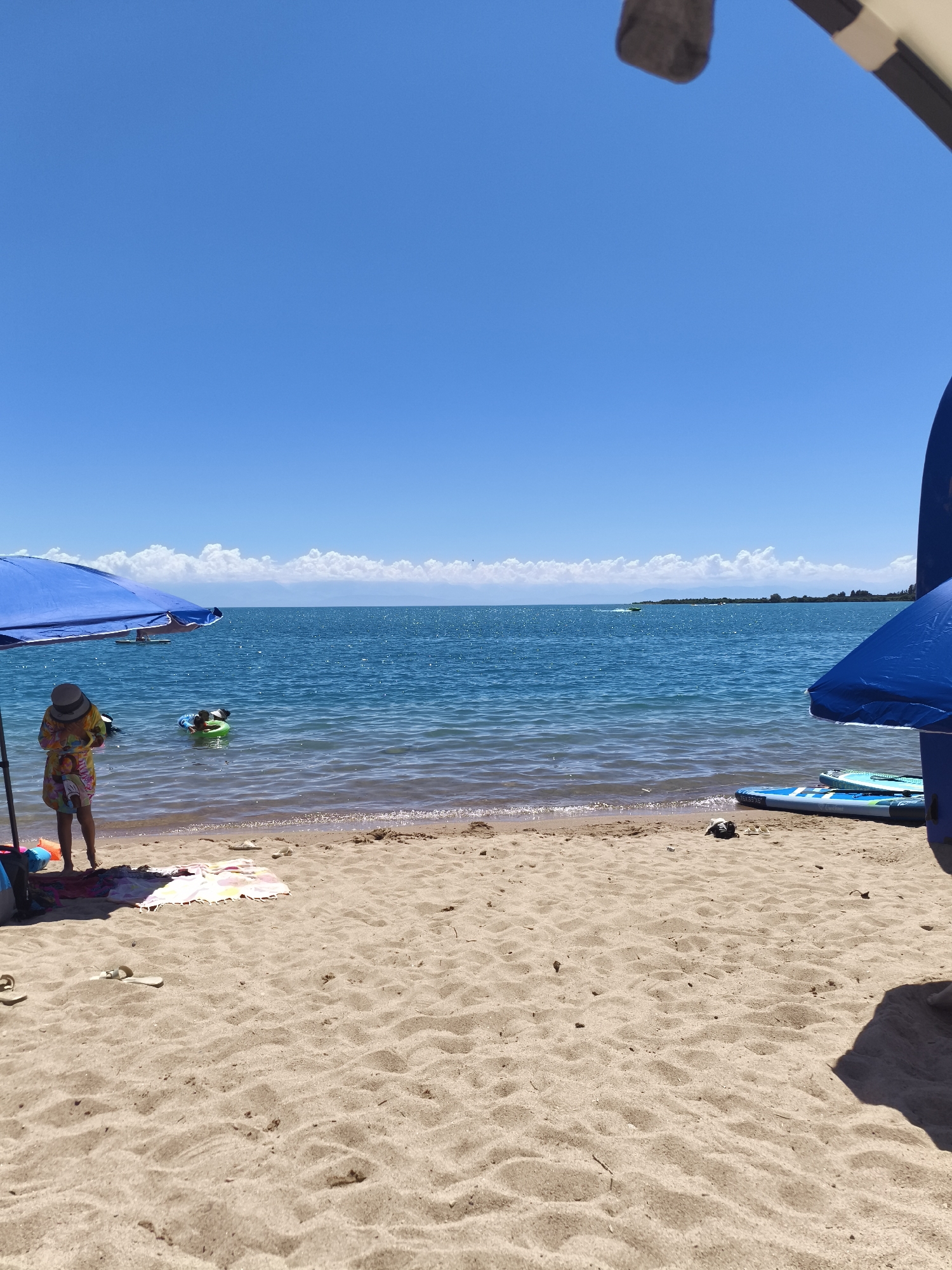 A sunny day at the beach in Bayet, Kyrgyzstan, is captured in this image. The foreground is dominated by sand, with a few flip-flops and a towel suggesting recent activity. A blue umbrella provides shade to a person wearing a straw hat and a colorful patterned dress, standing near the water's edge. To their left, a blue bag and orange flotation device are visible. In the water, a person is visible in a green flotation ring. Further out, the clear blue water meets a distant horizon line where scattered white clouds hover above a landmass. Several paddleboards are arranged on the sand to the right of the frame, with two blue umbrellas also present. The overall scene depicts a leisurely day at the lake.