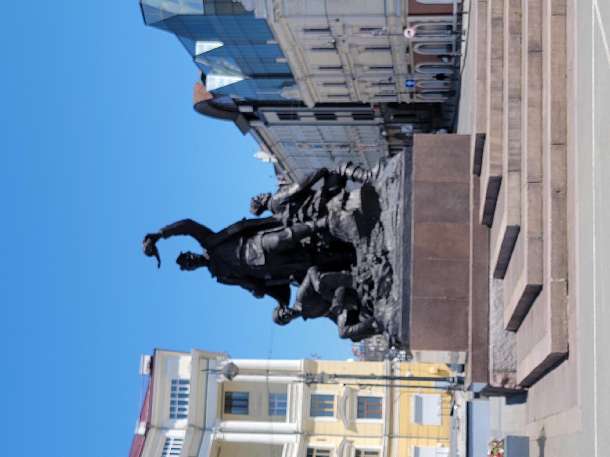 Here is a detailed description of the image:
The image captures a statue group in an urban environment, likely a monument within a city. The statues, cast in a dark material like bronze or a dark stone, depict figures engaged in what appears to be a conflict or a struggle. One figure, notably, is standing and gesturing dramatically, while others seem to be supporting him or involved in the action happening at ground level.
The statue group is mounted on a structure made of dark brown or reddish-brown stone, which serves as its base. Adjacent to this base, there is a stepped structure of a lighter-colored stone, possibly granite or a similar material, which could be part of a plaza or memorial.
In the background, there are buildings with diverse architectural styles. One building features a modern glass facade, while others exhibit more classical designs with multiple stories and ornate details. The sky above is clear and blue, suggesting it is daytime.
The location is Unknown, Unknown.