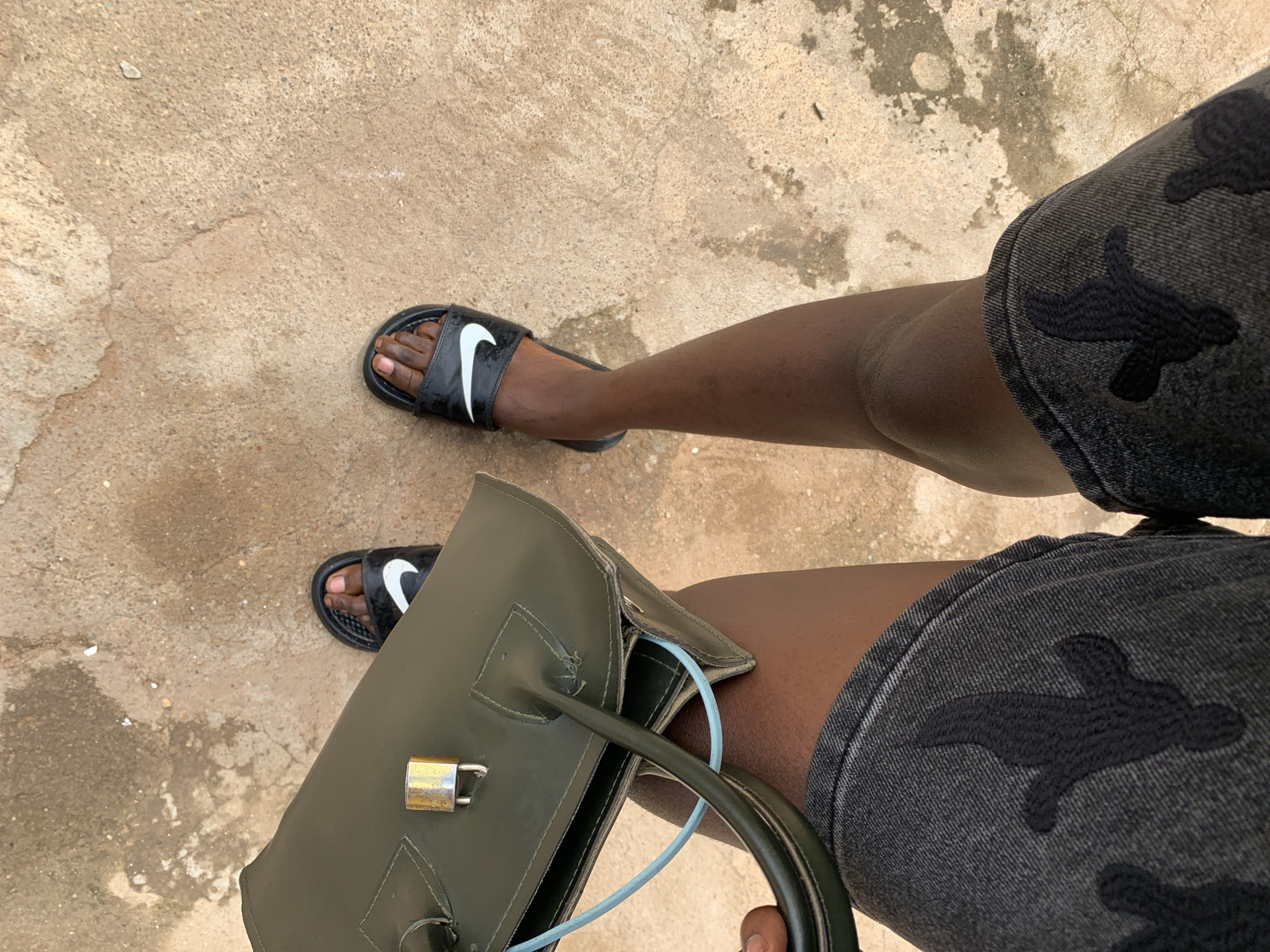 An overhead view captures the lower body of an individual standing on an irregular, light brown ground surface in Rafin Par, Nigeria. The person, exhibiting a dark complexion, is wearing dark grey denim shorts with a dark, abstract pattern. Both feet are clad in black Nike slide sandals, each featuring a white swoosh logo. The individual's right hand holds a dark olive-green structured handbag. A gold-colored padlock is visible on the front flap of the handbag, and a light blue cord or strap is partially visible within its opening. The ground exhibits a textured, unpaved, or roughly paved surface with variations in color and consistency.