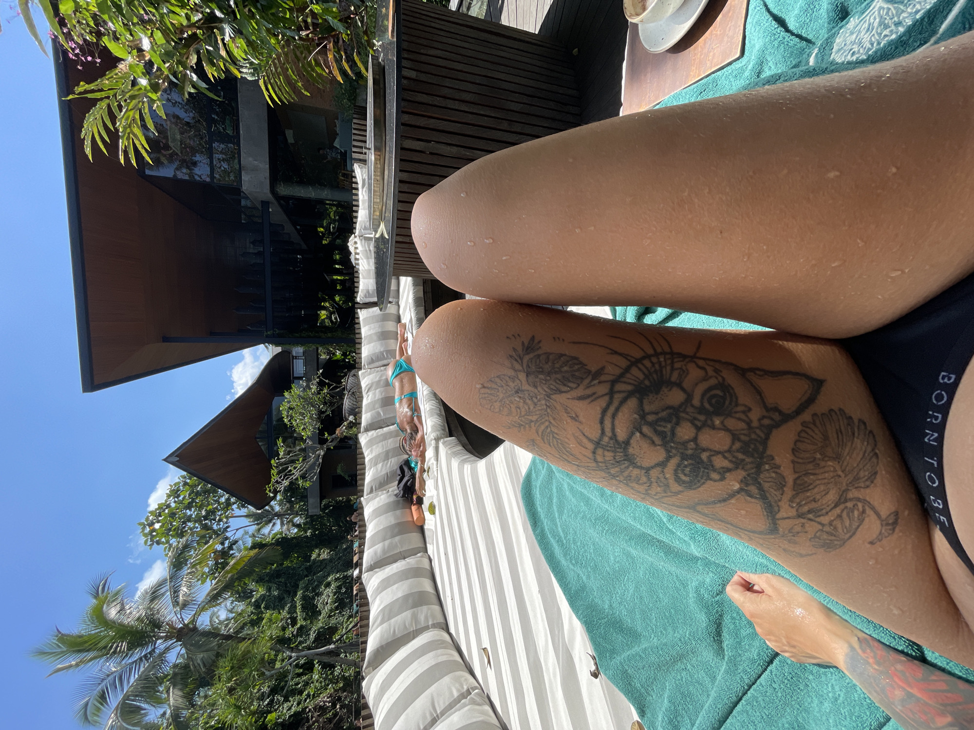 In Ubud District, Indonesia, on a sun-drenched day, two bare legs are prominently featured in the foreground, resting on a teal towel. The left leg displays a detailed black tattoo of a floral design and an animal face. A hand is visible to the left, resting on the towel. In the midground, a curved, white-striped sofa is occupied by several individuals lounging and sunbathing. One person is wearing a blue bikini. Behind the sofa, a modern, multi-level building with large windows and a wooden roof blends into the lush green foliage and palm trees that surround the property. The environment suggests a resort or private villa setting, with an outdoor wooden deck and a circular table visible in the background. The sky is a clear, bright blue with a few white clouds.