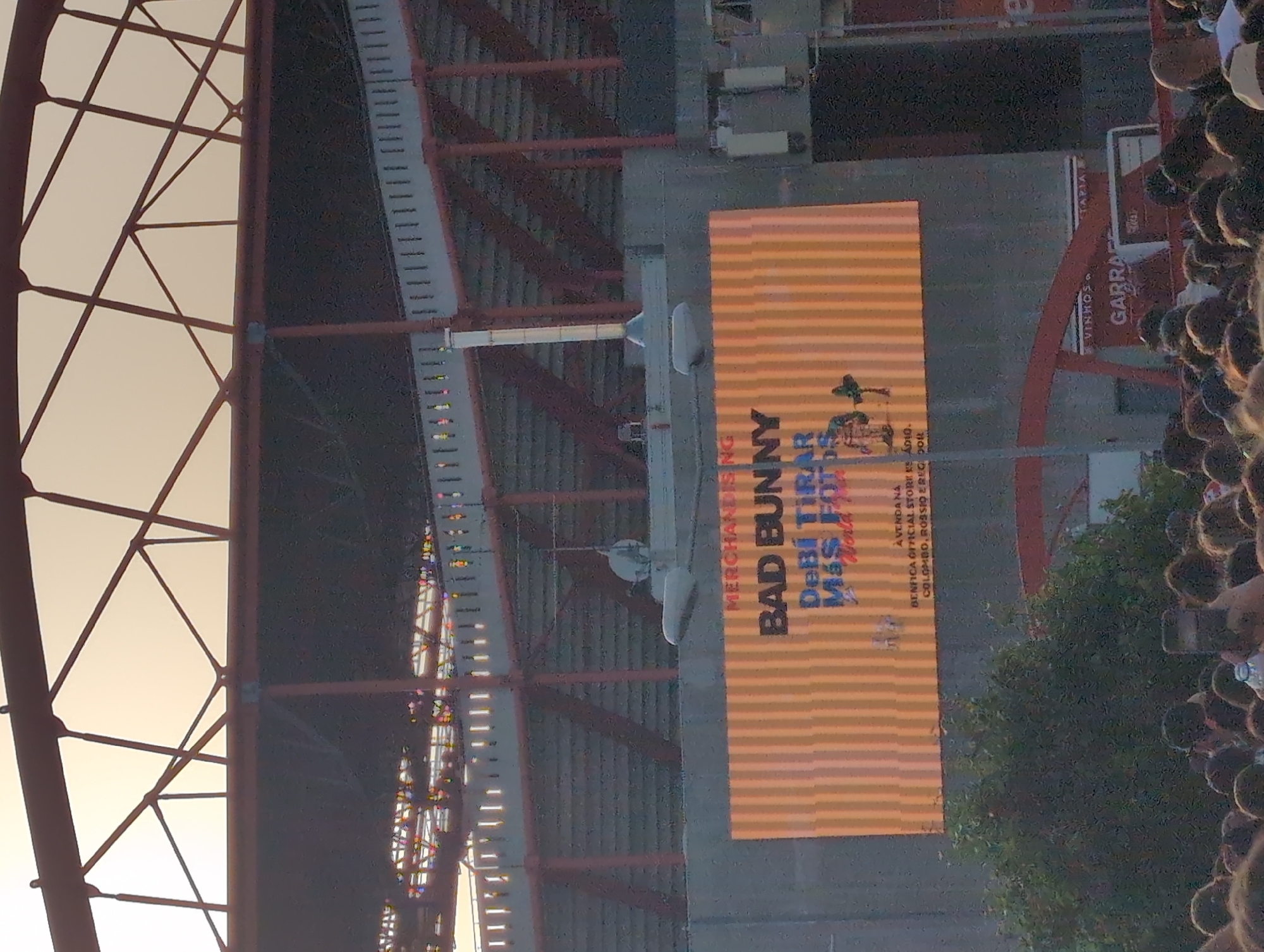 The image depicts an outdoor scene, likely near Estádio da Luz in Lisbon, Portugal, as indicated by the presence of a "BENFICA OFFICIAL STORE" advertisement. A large billboard advertises Bad Bunny merchandise, specifically mentioning "DeBi TIRAR M&S FOTOS." The billboard is mounted on a structure, possibly part of the stadium's exterior or a nearby building.
A crowd of people is visible to the right, suggesting a gathering or event related to the advertisement. A tree partially obscures the view of the crowd and the advertisement. Architectural details, including red beams and structural elements, are also visible, presumably related to the stadium or the surrounding infrastructure.
