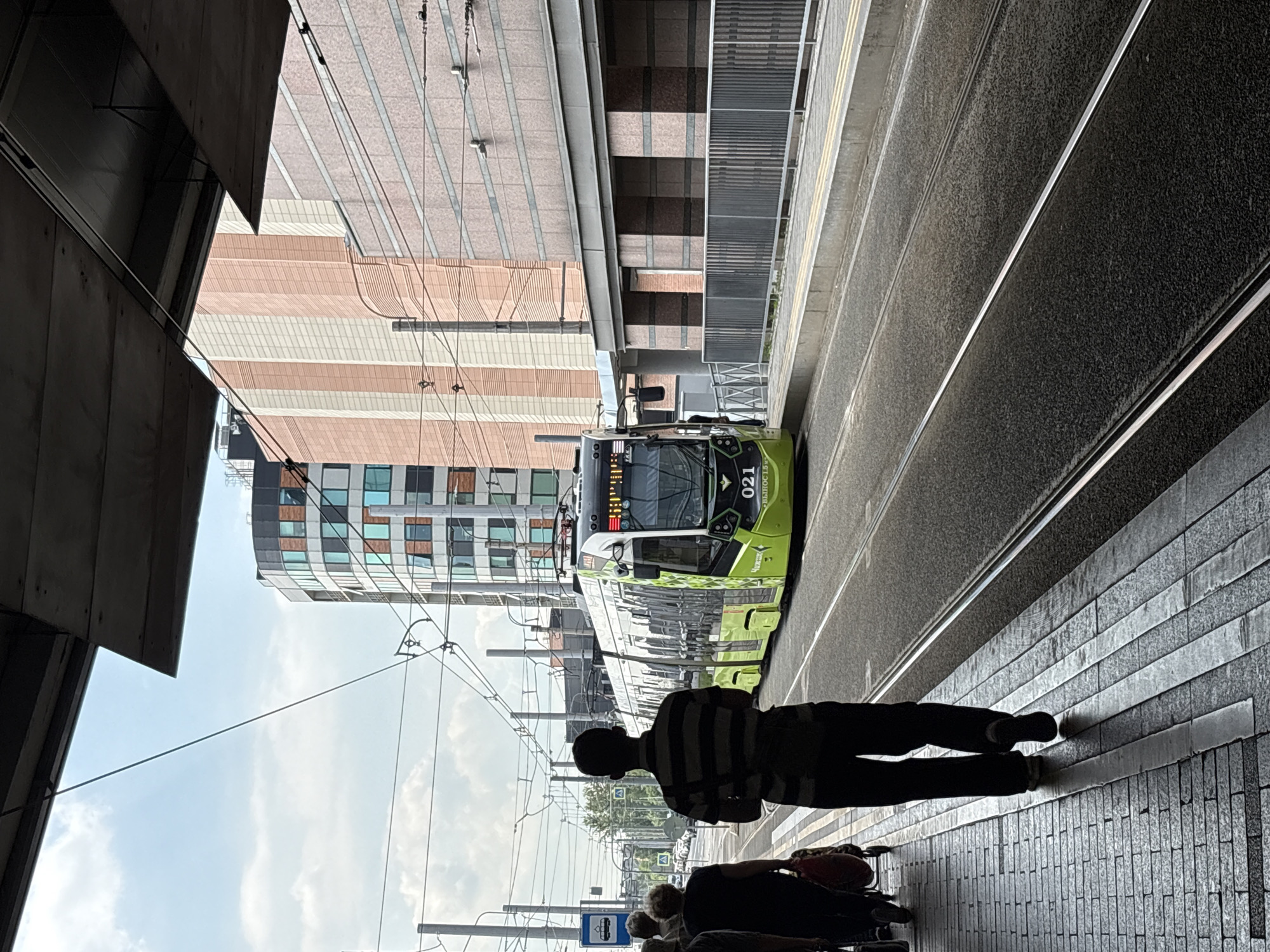 In Saint Petersburg, Russia, a bright green tram, numbered 021, is stopped at a tram station beneath an overhead structure. The tram has "TRM" written on its side. Several people are visible at the station. In the foreground, a person wearing a striped shirt and dark pants walks away from the camera on a brick sidewalk. To their left, other pedestrians, including a woman with a red bag, are also waiting. In the background, modern buildings with colorful facades and windows rise above the station. The tram tracks run parallel to the station platform and extend into the distance. The sky is partly cloudy.