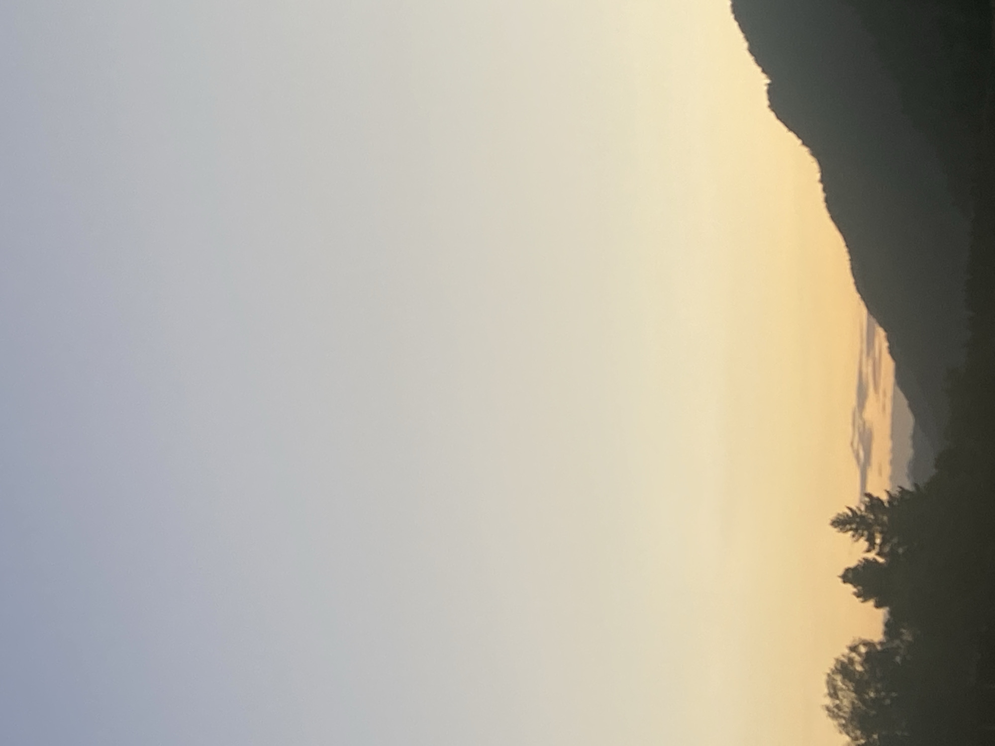 The image depicts a landscape scene with a gradient sky transitioning from a light blue at the top to a soft yellow near the horizon. Silhouetted against the sky are the dark outlines of trees and mountains. The foreground features several trees, two of which are prominent evergreens. In the middle ground, a range of hills or mountains creates a jagged silhouette. The background shows the faint contours of distant mountains under the twilight sky. The overall impression is one of a serene natural environment at dusk or dawn. The provided crops focus on sections of the sky and the silhouetted landscape, offering closer views of the color gradients and the texture of the mountains. There are no people or discernible man-made structures visible in the image. The scene is located in the city of Dzhambul, Kazakhstan.