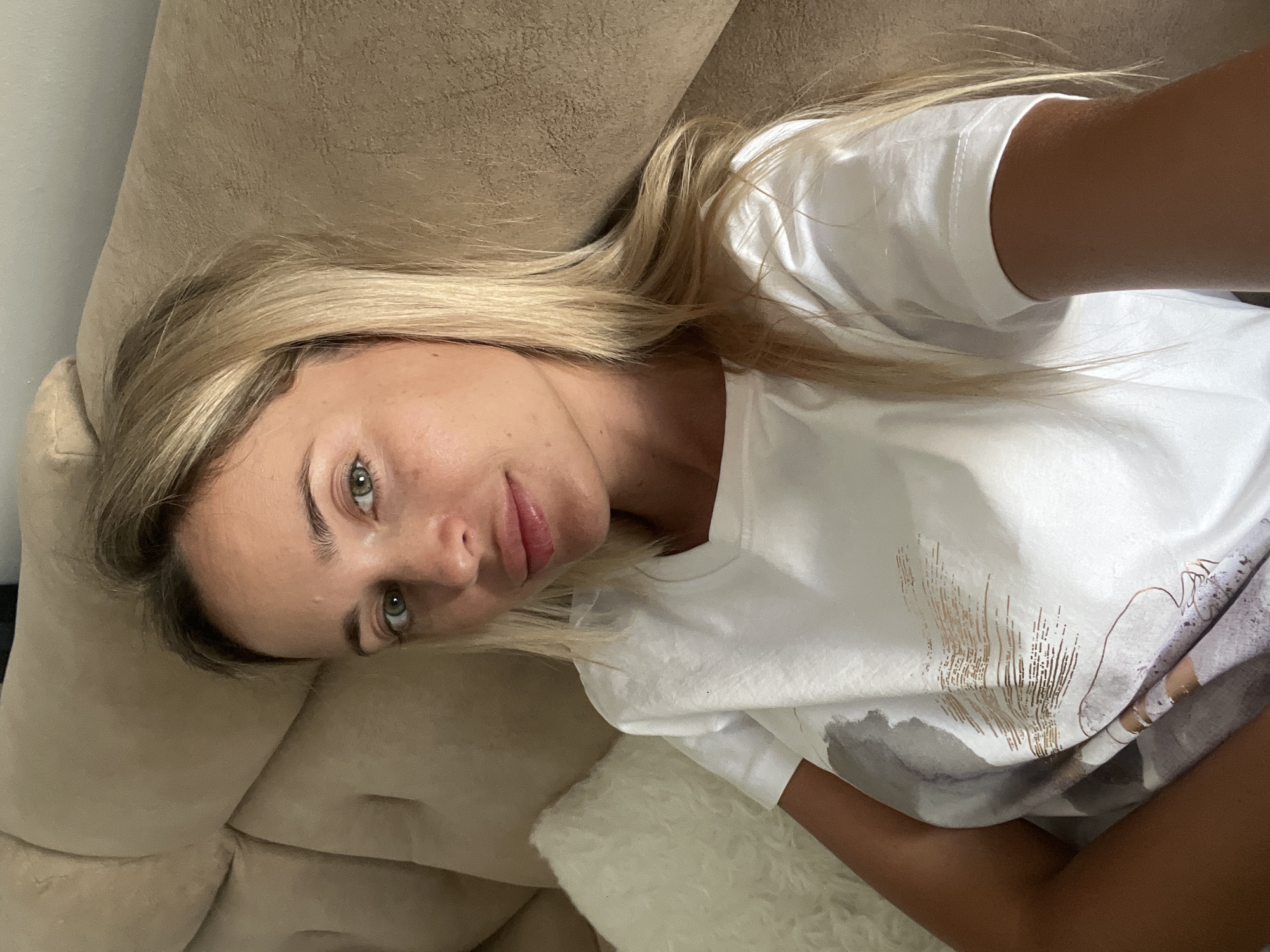 In Sharm El Sheikh, Egypt, a young woman with blonde hair and green eyes is captured in a selfie, leaning back on a beige couch. She is wearing a white t-shirt with a subtle abstract pattern. A white, fluffy pillow is visible to her left. The lighting suggests an indoor setting, likely a living room.