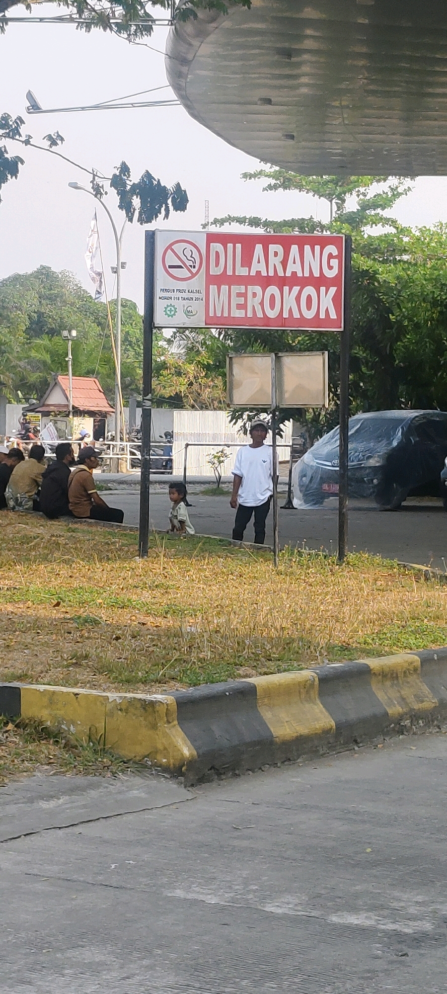 In Banjarmasin, Indonesia, a "No Smoking" sign, written in red and white, is prominently displayed on a metal pole. Below the main "DILARANG MEROKOK" (No Smoking) text, smaller writing indicates "PERGUB PROV. KALSEL NOMOR 018 TAHUN 2014," along with a green logo featuring a medical symbol and "MA". Two empty, framed panels are attached below the sign.
In the foreground, a child with dark hair, wearing a light-colored outfit, sits on the grass, facing towards the sign. To the right of the child, a man in a white t-shirt and dark pants stands, looking towards the right side of the frame.
In the background, to the left, a group of people are seated on the ground, some appearing to be resting. A small building with a red roof is visible behind them, along with a street light. A black car, partially covered with a tarp, is parked to the right of the main sign. The environment appears to be an outdoor area with dry grass and trees, possibly a roadside or a public waiting area, with a concrete barrier in the immediate foreground.