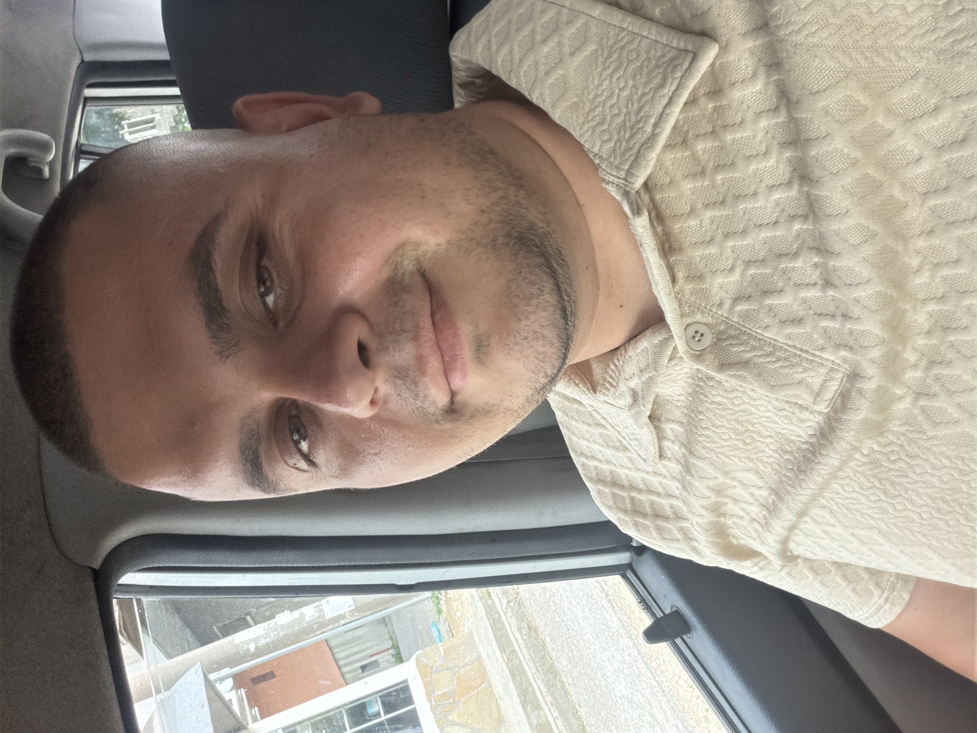 A male individual is depicted in a head-and-shoulders shot, seated inside a vehicle. He has fair skin, a rounded face, dark brown eyes, and dark, closely-cropped hair with a buzzed fade on the sides. Short stubble is visible on his jawline and chin. He maintains direct eye contact with the camera and displays a slight, closed-mouth smile. He is wearing a light beige polo shirt with a discernible textured diamond pattern, featuring a visible collar and one unbuttoned top button.
The interior of the vehicle is visible, including a dark grey door panel with a window on the left, and a dark grey headrest and a glimpse of a rear window on the right. Through the left window, an exterior scene is visible, featuring part of a building with a white-framed window and a light-colored, possibly stone or concrete, foundation. Behind this, another structure with sections of orange-brown and grey is present. The ground outside appears paved or dusty. The rear window on the right offers a limited view of green foliage. There are no identifiable landmarks or specific architectural features within the image to confirm the location as Shumen, Bulgaria.