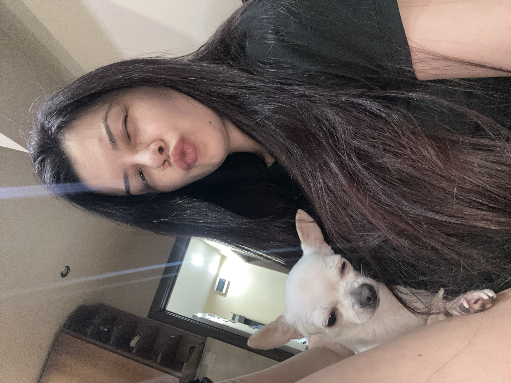 A photograph shows a woman and a dog in an indoor setting. The woman has dark hair and is wearing a black shirt. She is looking at the camera with her lips pursed. A small, light-colored dog is nestled beside her, partially obscured by her hair. The background includes a door frame, a light fixture, and a piece of furniture. There is no information provided about the location being Sliven, Bulgaria.