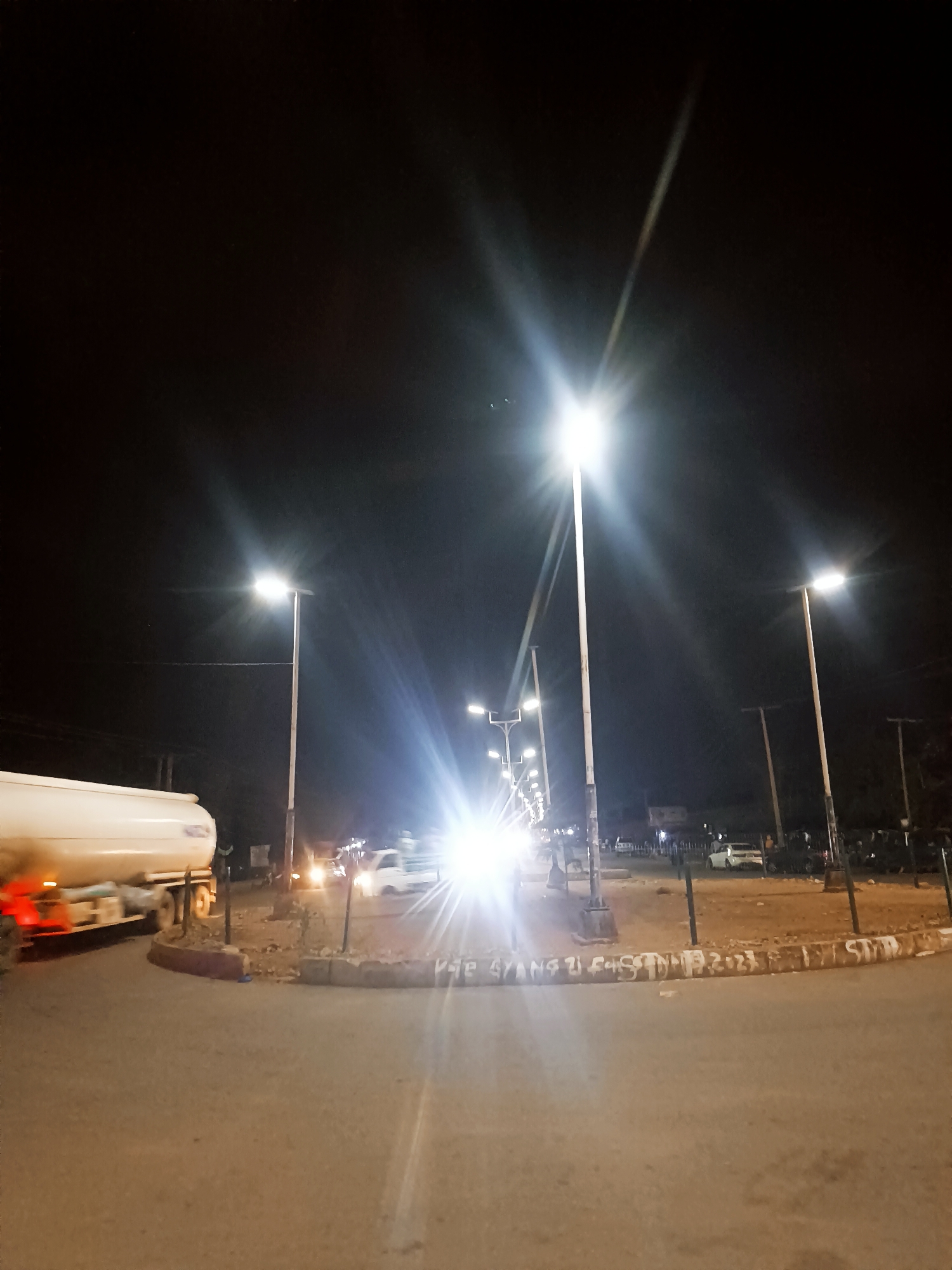 The image depicts a night scene in Bukuru, Nigeria. The location appears to be a roundabout or traffic circle illuminated by bright streetlights. Several lampposts line the road, casting strong light flares. A tanker truck is positioned on the left side of the frame, partially visible. There are additional cars in the distance, their headlights adding to the light sources. In the background, buildings and trees are barely visible against the night sky. The road surface is visible in the foreground, and the edge of the roundabout is lined with a low, concrete barrier covered with graffiti. Some blurry figures are also visible in the scene.