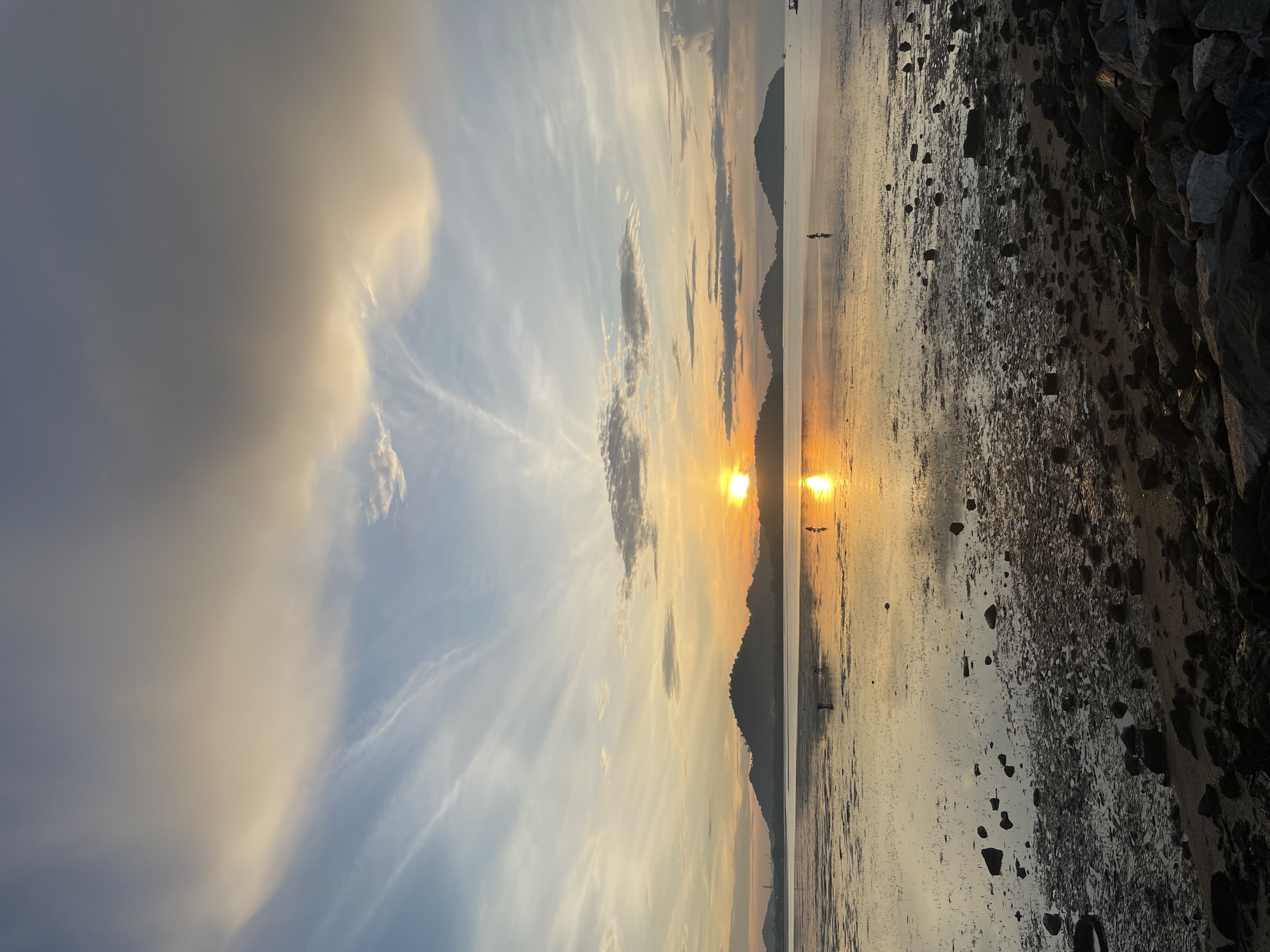 The image captures a serene sunset over a coastal area in Phuket City Municipality, Thailand. The sun, a bright orb of golden light, is low on the horizon, casting a warm, reflective glow across the calm water. Several silhouetted figures, appearing to be individuals, are visible in the shallow water, some standing and others appearing to be in motion, possibly walking or fishing. The foreground of the image is dominated by a rocky shoreline, with stones of varying sizes scattered across the wet sand. In the distance, a chain of dark, tree-covered hills forms a silhouette against the colorful sky. The sky itself is a canvas of blended colors, with streaks of pale orange and yellow from the setting sun mingling with soft blues and grays, punctuated by wispy clouds. The overall scene evokes a sense of peace and natural beauty at dusk.