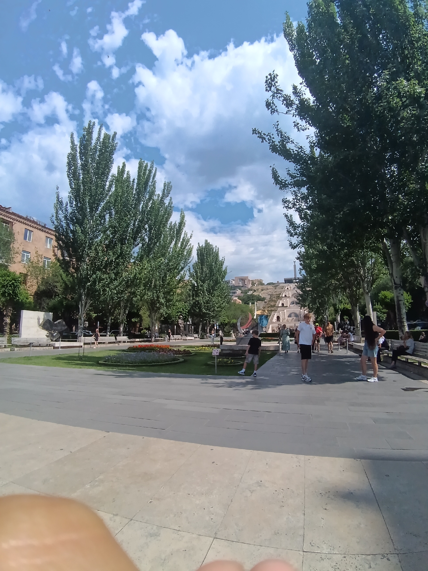 The image depicts a wide shot of a public plaza on a sunny day. Tall trees with lush green foliage line a paved walkway where several people are strolling. In the background, a large, stepped structure, possibly a cascade or stairway, is visible, with buildings and more trees on a hillside beyond. The sky is blue with scattered white clouds, and the ground is paved with large, light-colored tiles. A flower bed with colorful blooms is situated on the left side of the plaza. People are dressed casually, and the overall atmosphere appears relaxed and lively.
