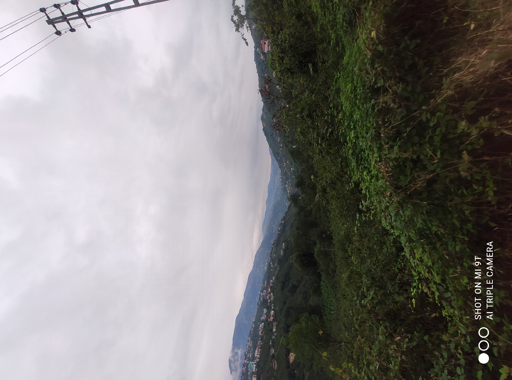 A wide shot captures a hazy, overcast day in Akçaabat, Turkey, overlooking a valley dotted with houses cascading down rolling hills. Dense green foliage dominates the foreground, with lush bushes and trees obscuring the lower half of the image. In the mid-ground, a settlement of white and red-roofed houses is visible, spread across the steep slopes of the mountains. A minaret of a mosque is identifiable among the buildings. The background is filled with more mountains, fading into the mist and clouds. A utility pole with wires is positioned on the right side of the frame, against the pale sky. The bottom right corner of the image displays the watermark "SHOT ON MI 9T AI TRIPLE CAMERA".
