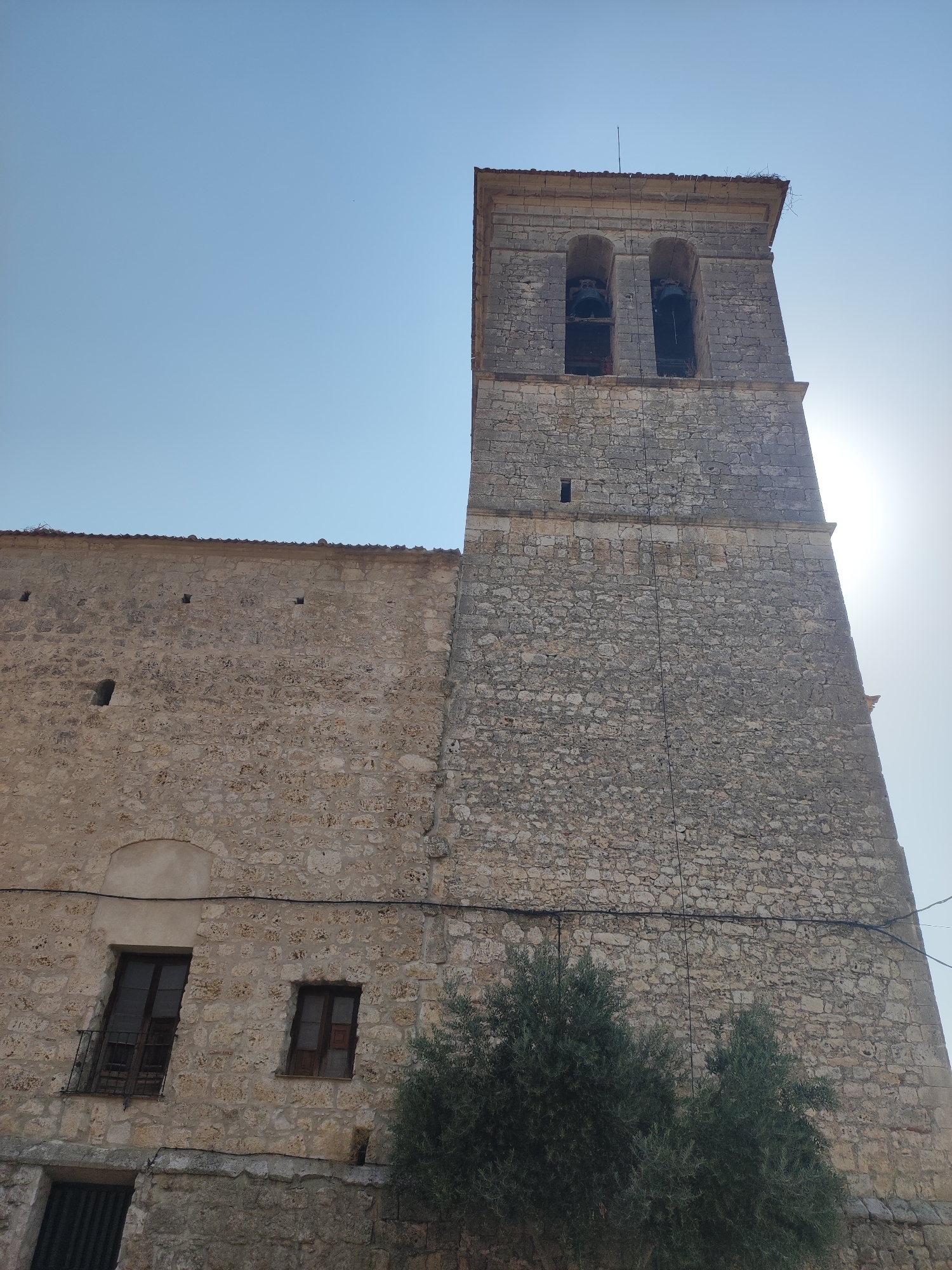A low-angle, full outdoor shot captures the facade of a stone church in Traspinedo, Spain. The sky is clear and pale blue. The church has a tall, square bell tower made of grey stone blocks. The tower has two arched openings at the top, each containing a bell. The main body of the church is made of lighter, rougher stone. There are several windows visible on the church walls, some with wooden frames and one with a small balcony. A small, green tree grows at the base of the church, partially obscuring the lower section. The overall impression is one of an old, weathered stone structure against a bright sky.