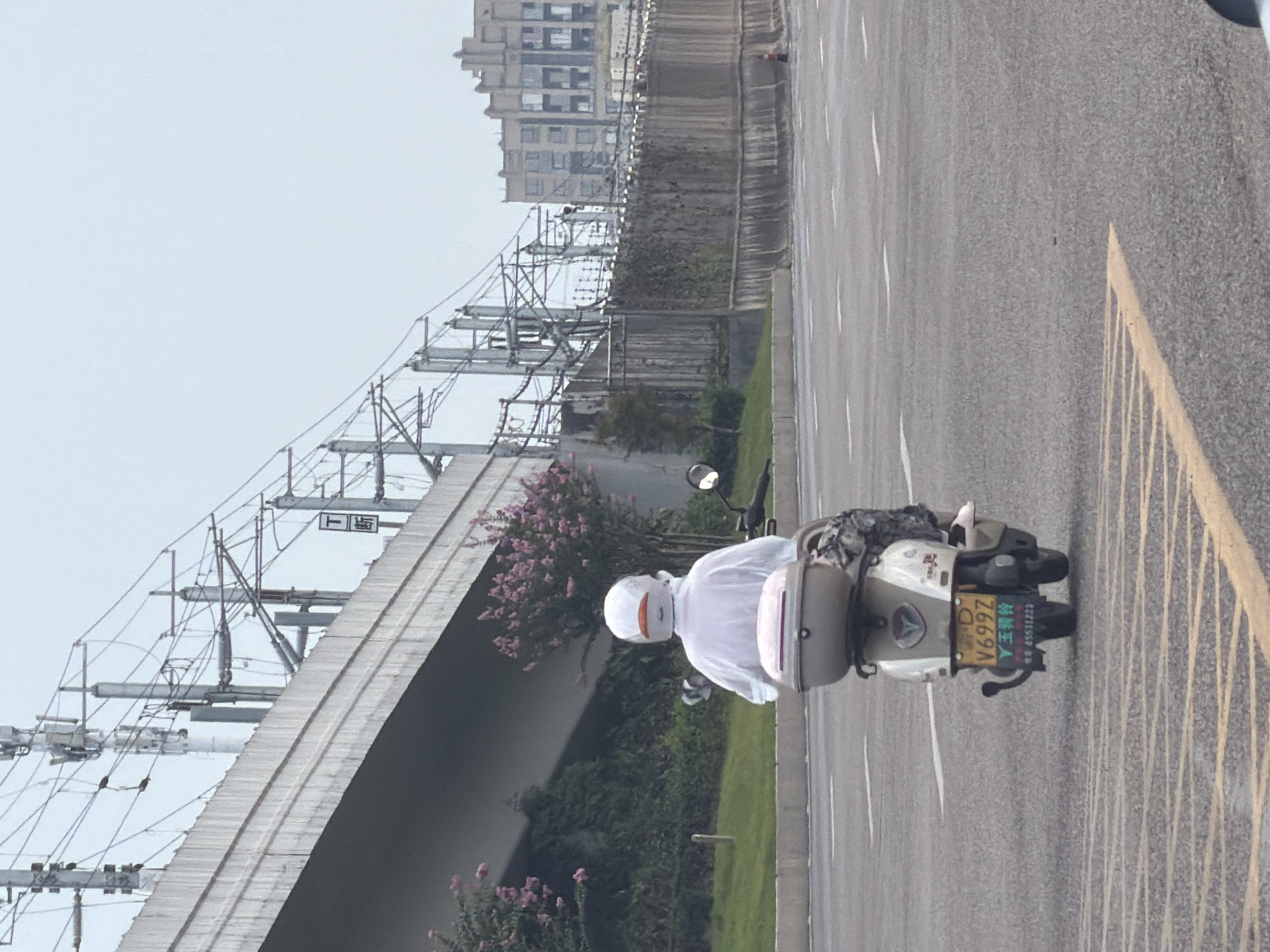 A person wearing a white helmet with an orange face decoration is riding a scooter on an asphalt road in Jiangjin, China. The scooter has a white and light brown body and a gray storage box on the back. The license plate reads "D V699Z". The person is wearing a white long-sleeved shirt and patterned leggings. In the background, there is an elevated road, power lines, trees with pink flowers, and buildings. The sky is overcast.