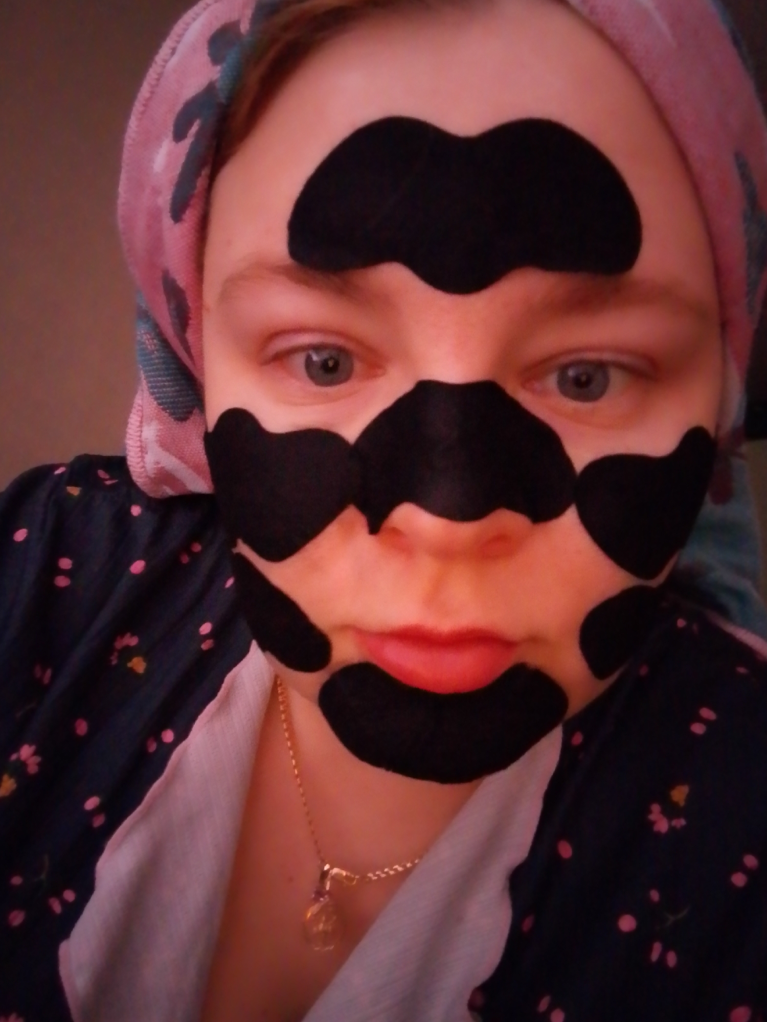 A fair-skinned person with blue eyes is shown from the chest up. They have sections of black peel-off mask applied to their forehead, under each eye, on the nose, and above the mouth. They wear a pink and blue patterned towel on their head and a dark blue shirt with small pink and yellow floral patterns over a light pink undershirt. A gold necklace is visible around the neck. The background is a solid, dark color, possibly a wall. There is no information to indicate this photo was taken in Izhevsk, Russia.