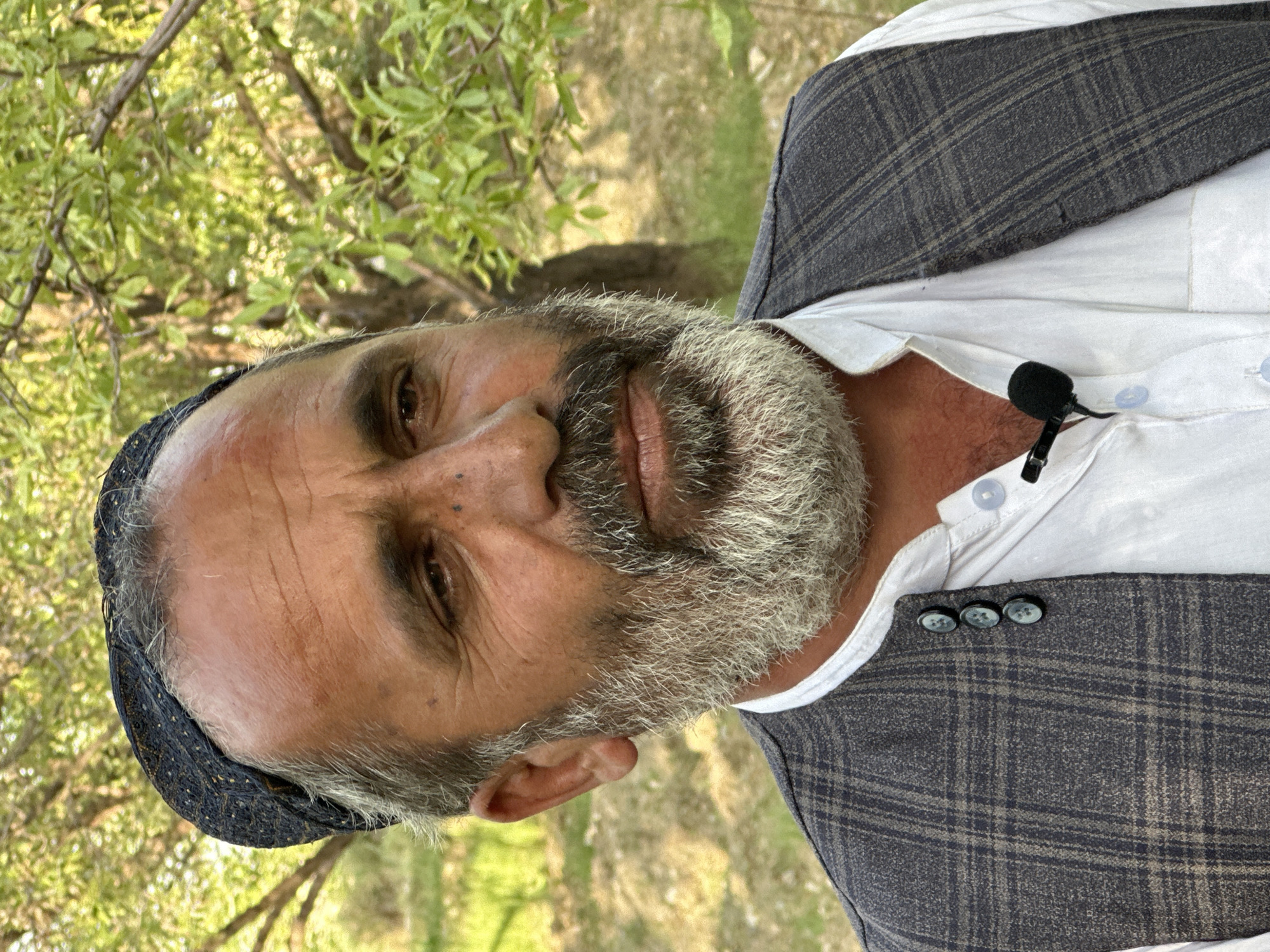 A middle-aged man with a gray beard and a dark cap is facing the camera. He is wearing a white collared shirt and a gray plaid vest. A small microphone is clipped to his shirt. The background is out of focus, but appears to be a grassy area with trees. The location is Unknown, Unknown.